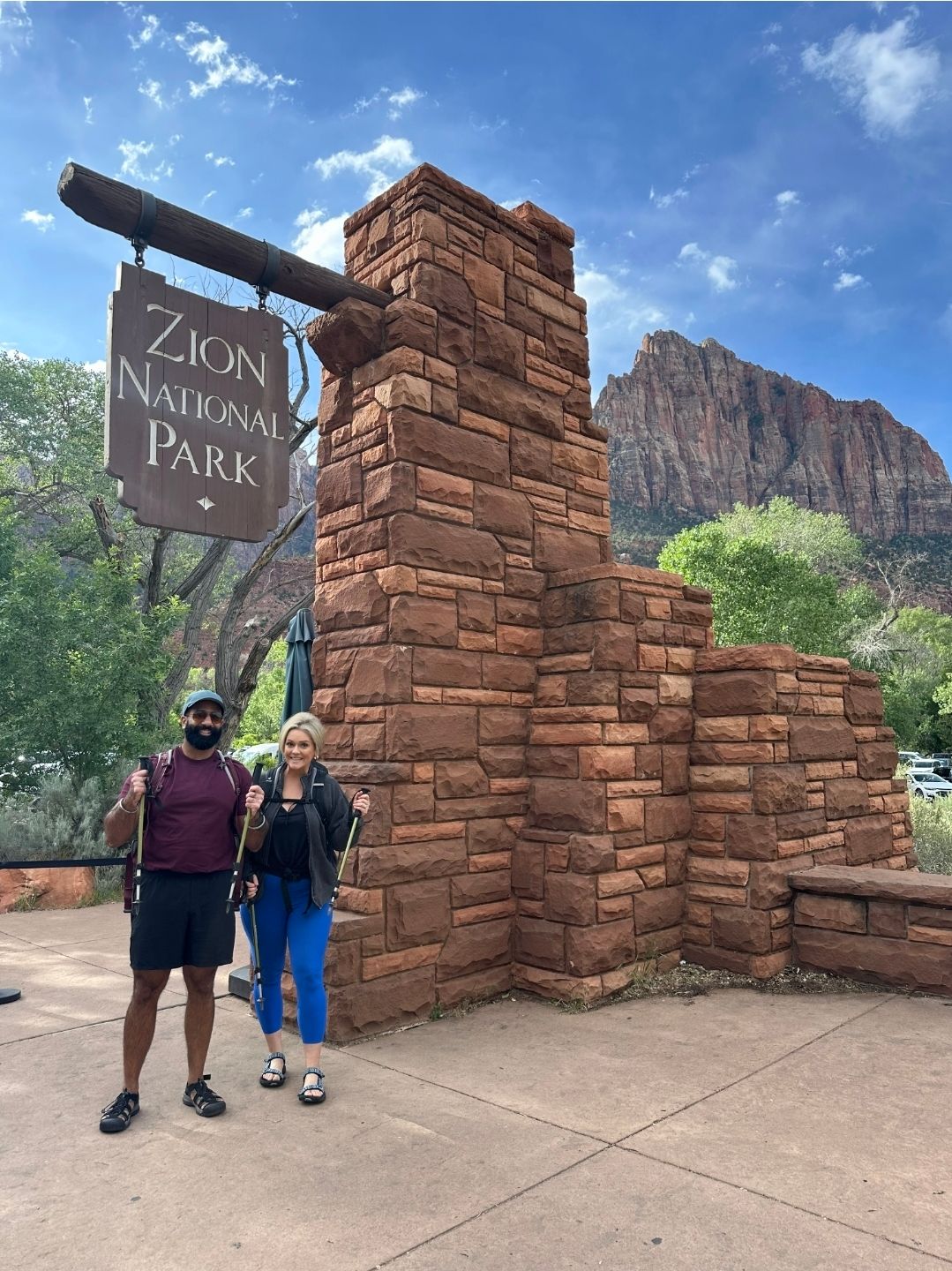 Couple posing at the Zion National Park entrance sign with red sandstone structure and mountain backdrop.