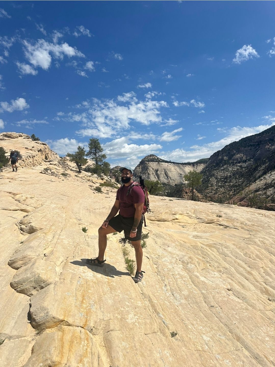 Person wearing a mask and backpack standing on sandstone in a sunny outdoor setting with mountains and blue sky.