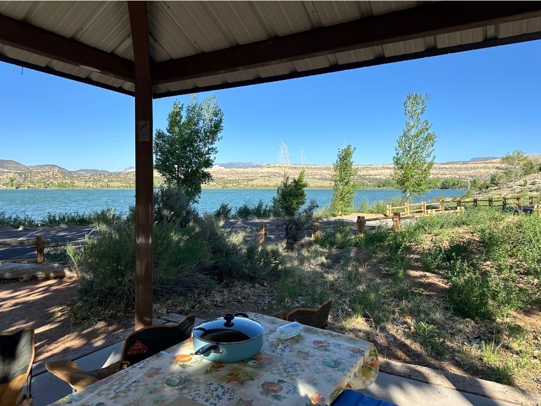 Picnic table under a shelter overlooking a lake and distant hills on a sunny day.