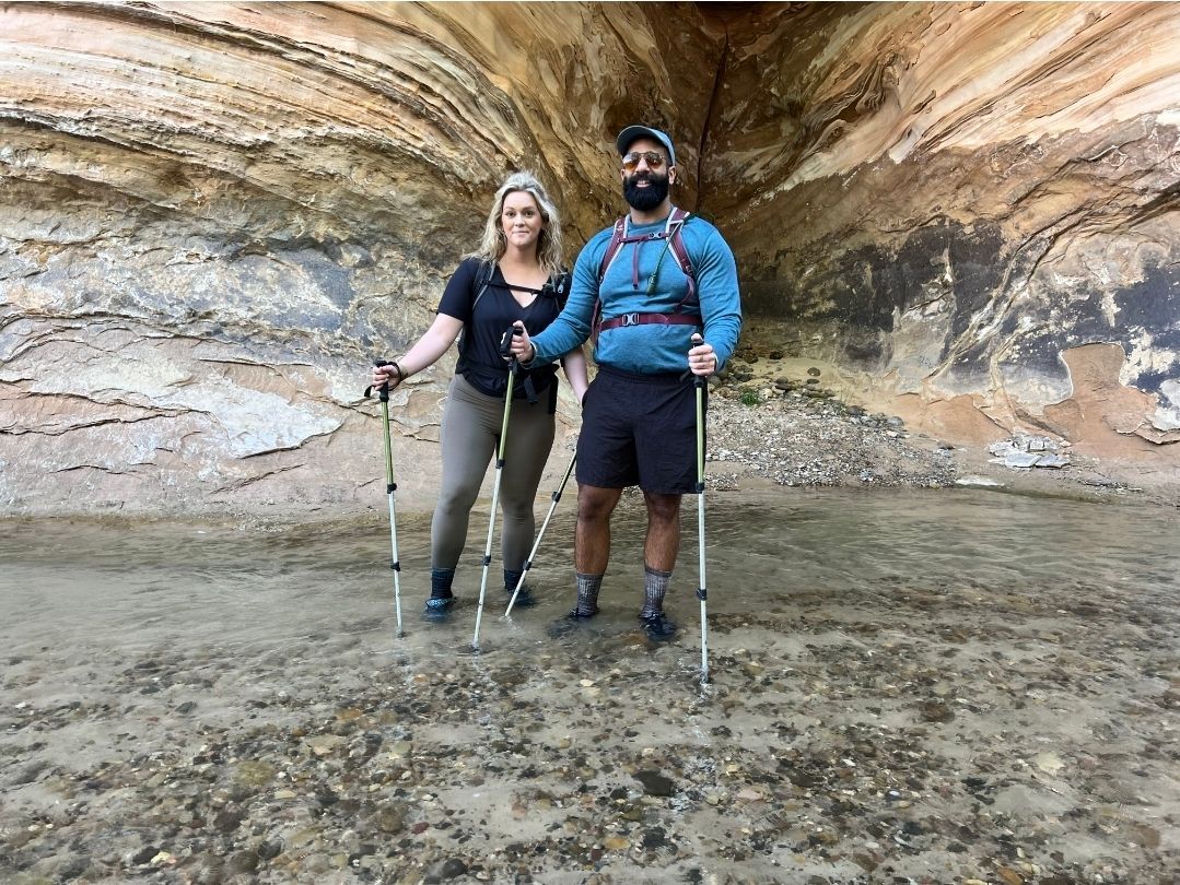 Two hikers holding hands under a rock overhang, using trekking poles. Background has layered rock formations.