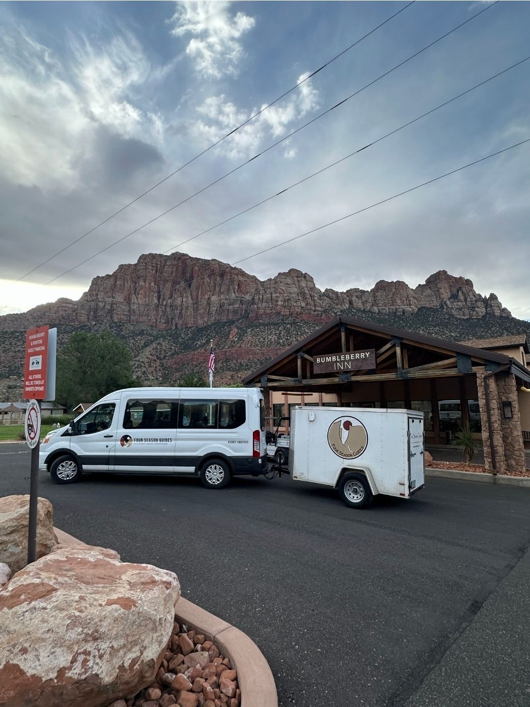 White van towing a trailer parked near a building with a mountainous backdrop. Cloudy sky.