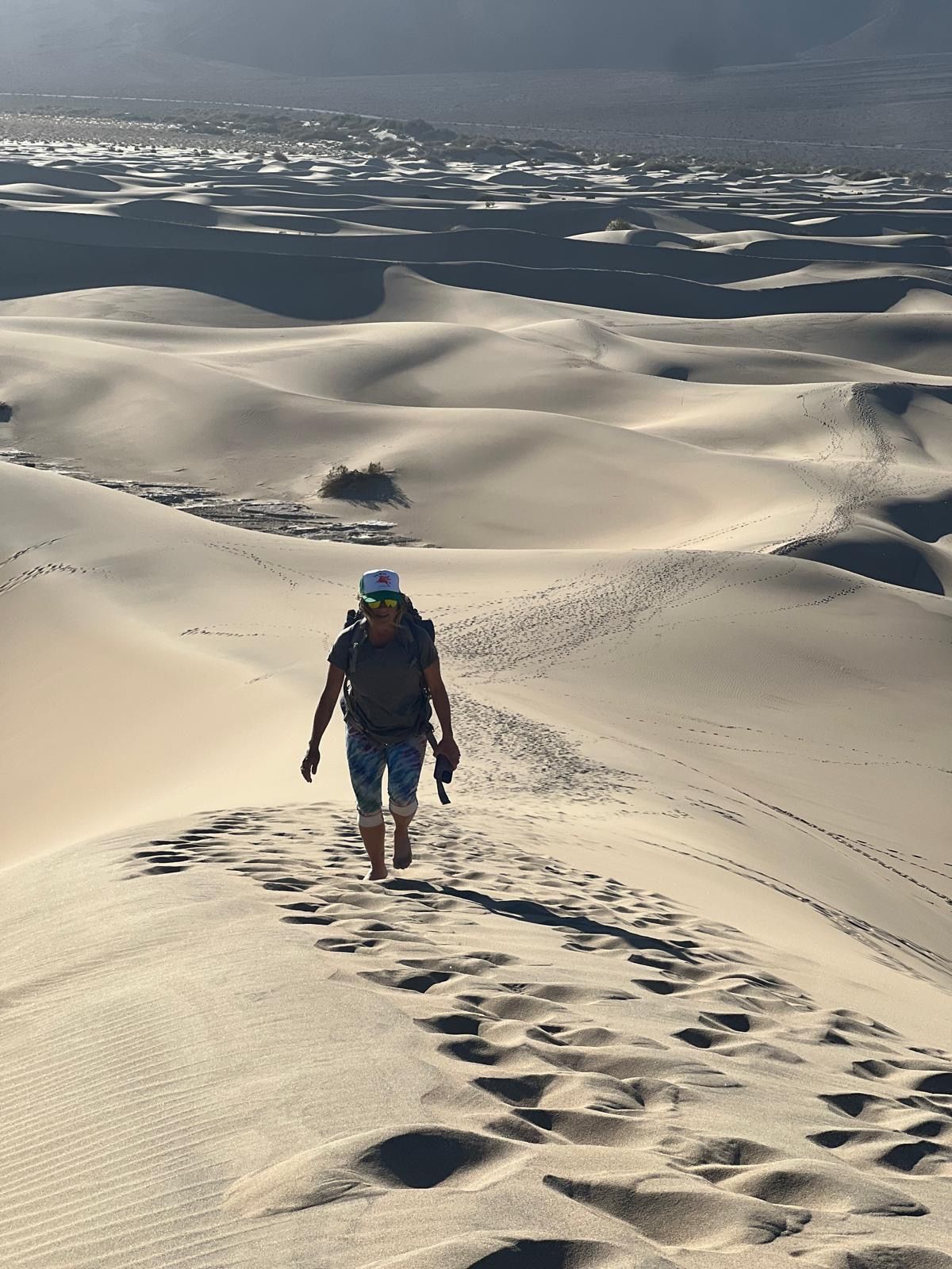 Person hikes across sand dunes, leaving footprints. Distant mountains and desert landscape visible.