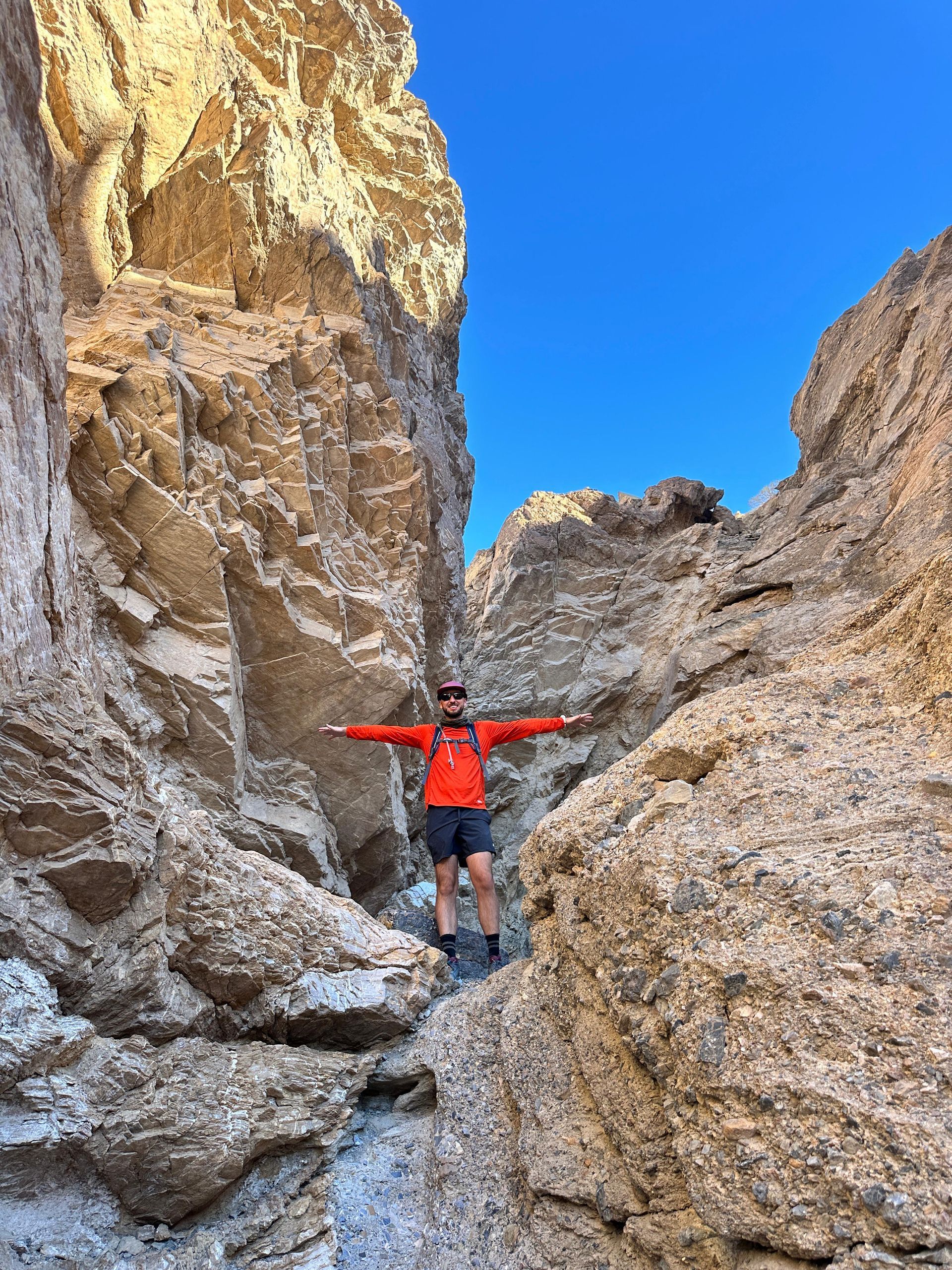 Person with arms outstretched in a narrow canyon, wearing orange jacket and shorts. Bright blue sky.