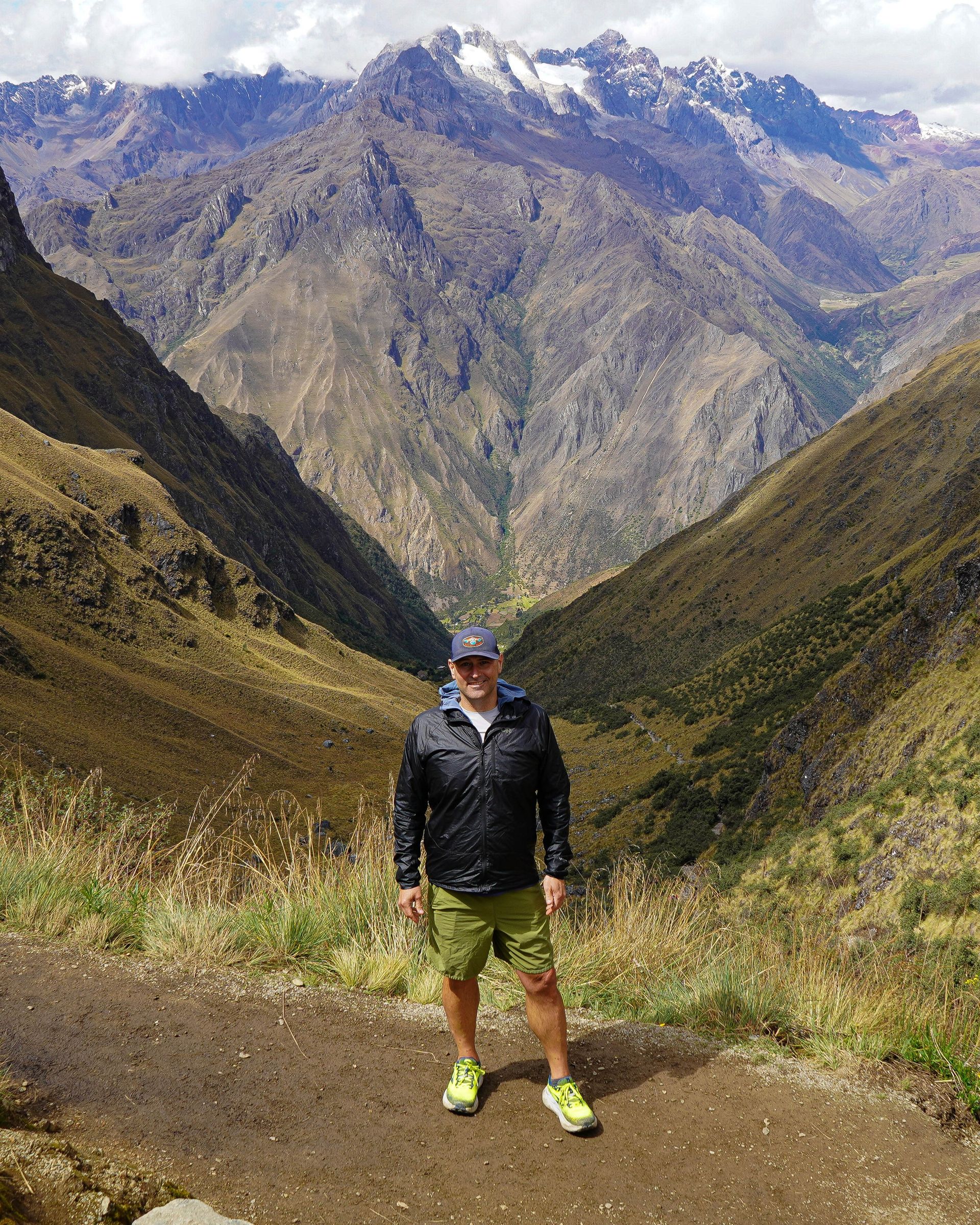 Man in shorts and jacket stands on a trail with mountain views.