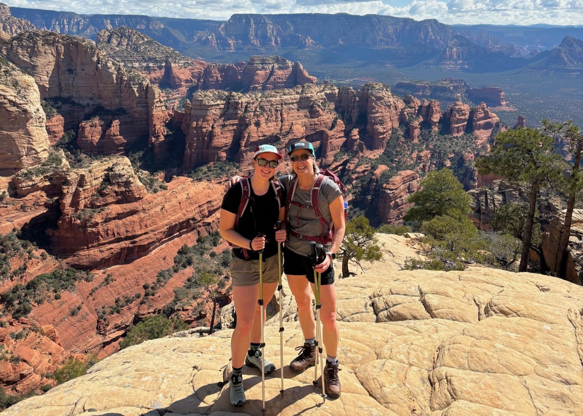 Five people stand on a large rock, arms raised, near a lake under a blue sky.