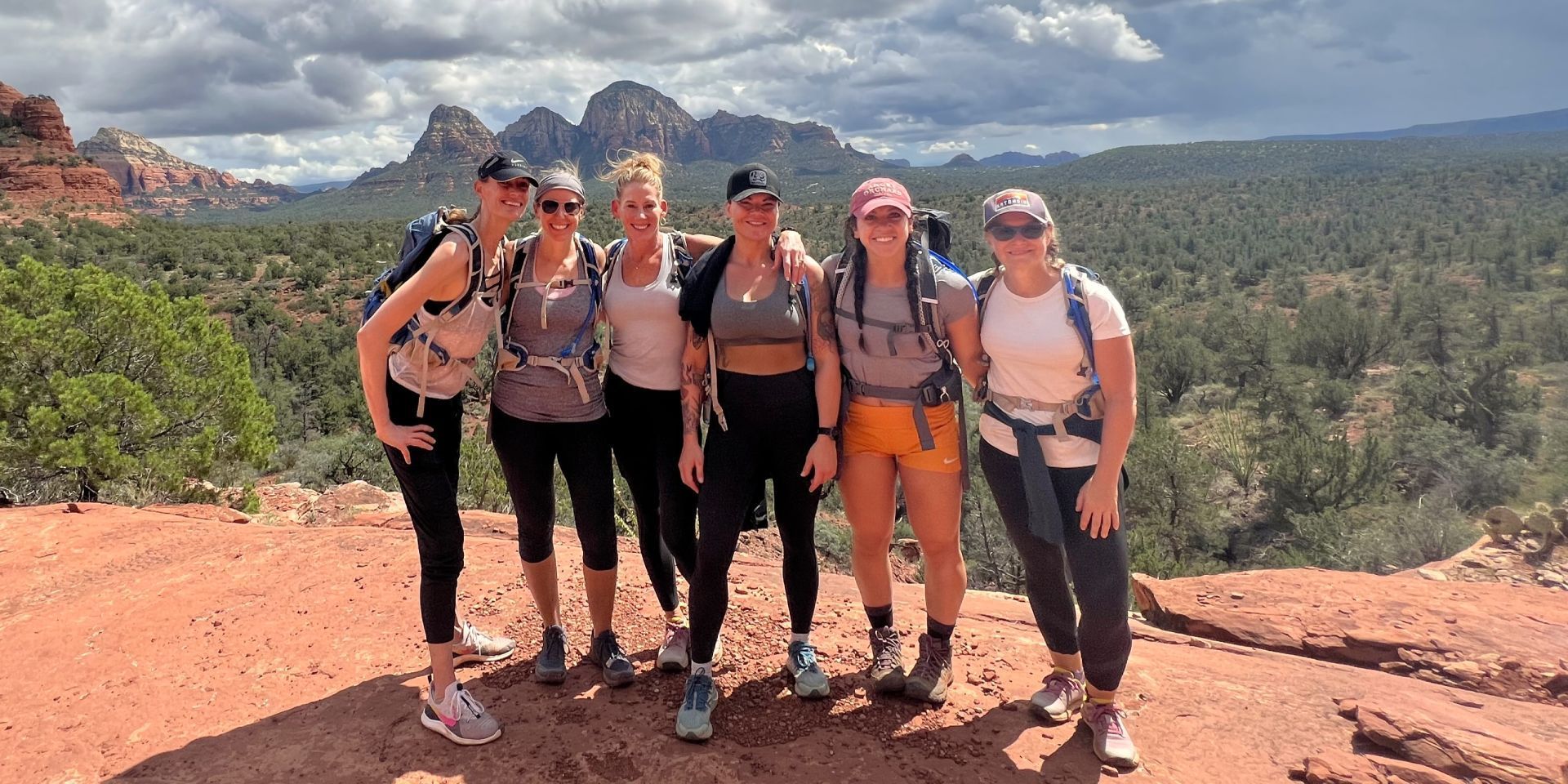 Seven women hikers posing on a red rock overlook, with a mountain backdrop and blue sky.
