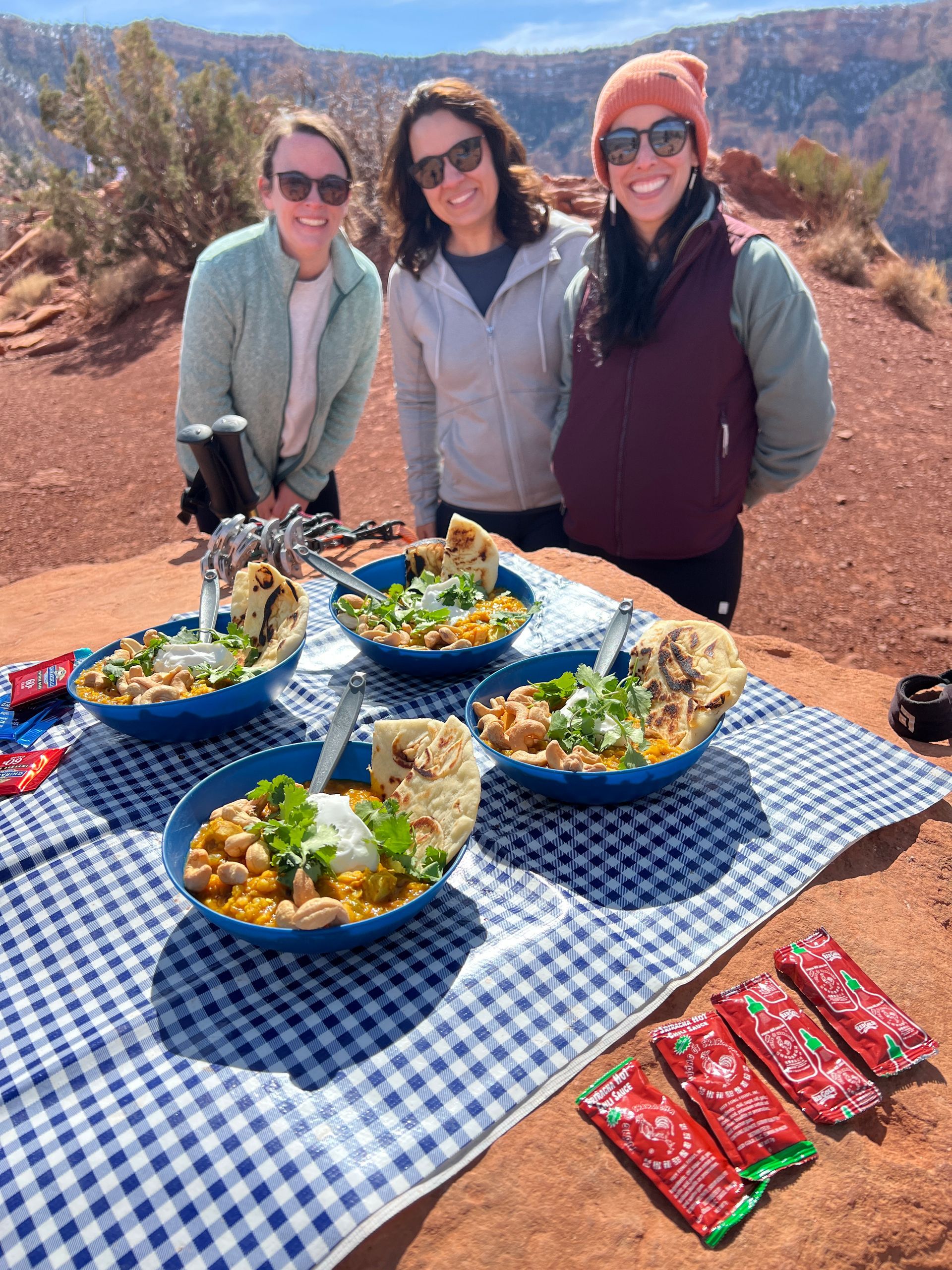 Three people smile near bowls of food on a picnic blanket, red rock backdrop.