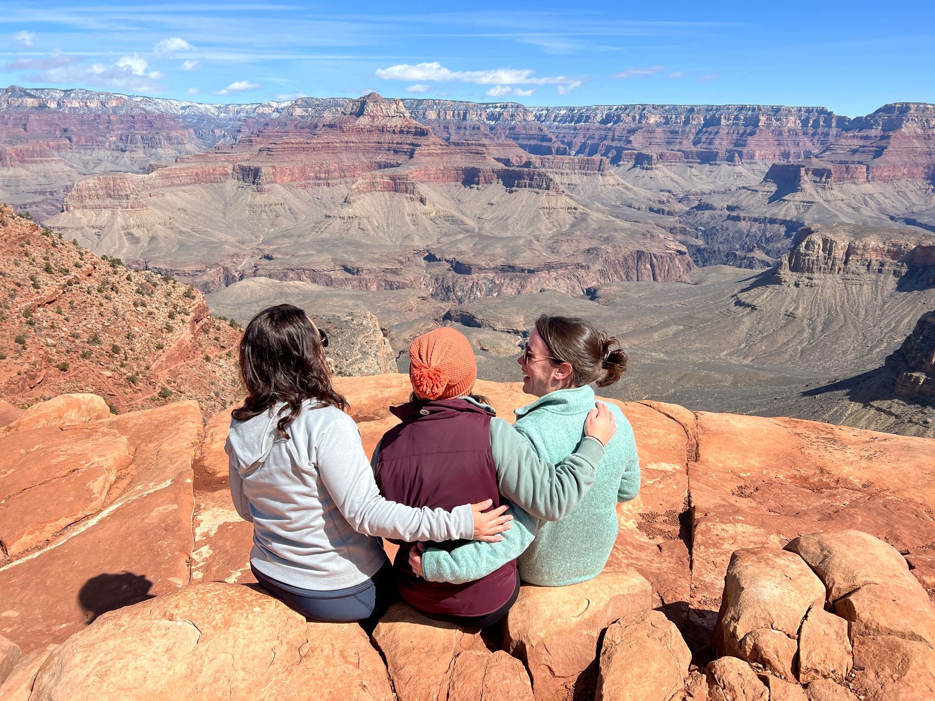 Three people sit on a rock, arms around each other, overlooking the Grand Canyon. Blue sky, red rocks.