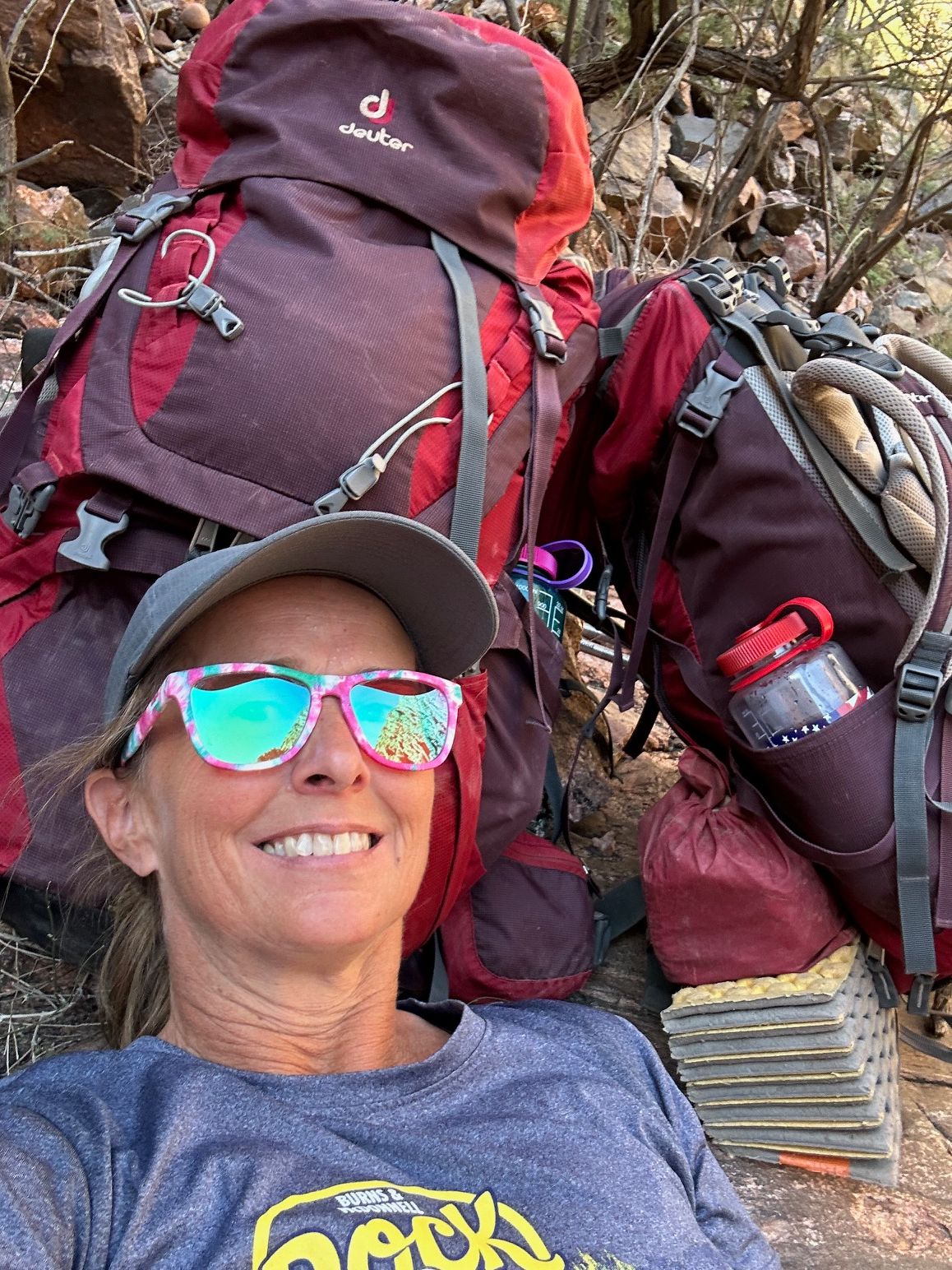 A woman wearing sunglasses and a hat is laying on the ground next to a backpack.