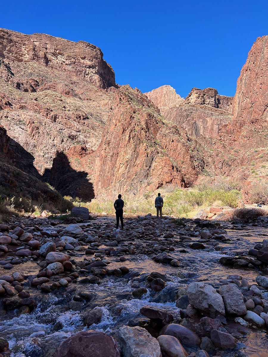 Two figures stand in a rocky creek bed, dwarfed by red canyon walls under a clear blue sky.