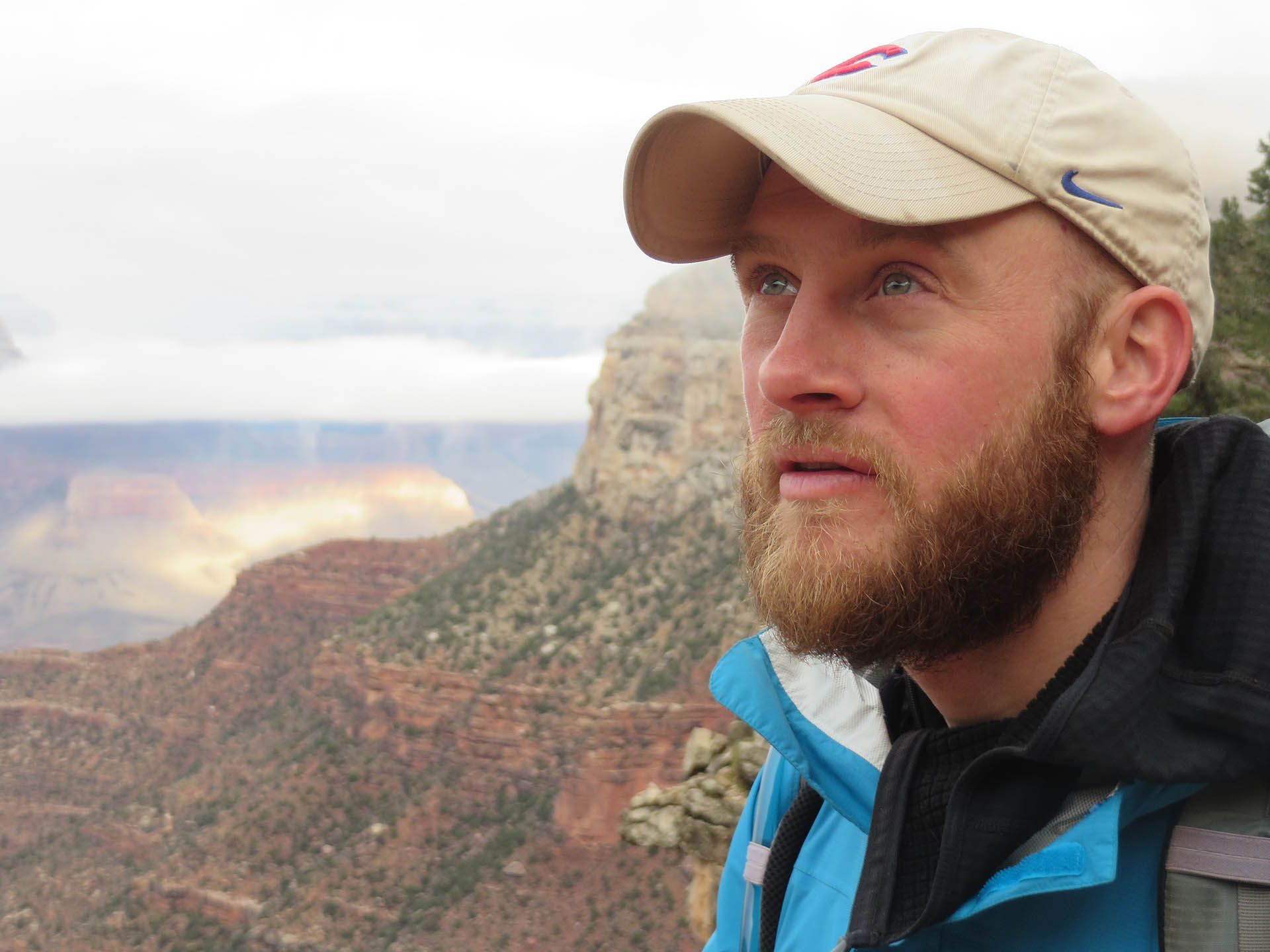 A man with a beard and a hat is standing in front of a mountain.