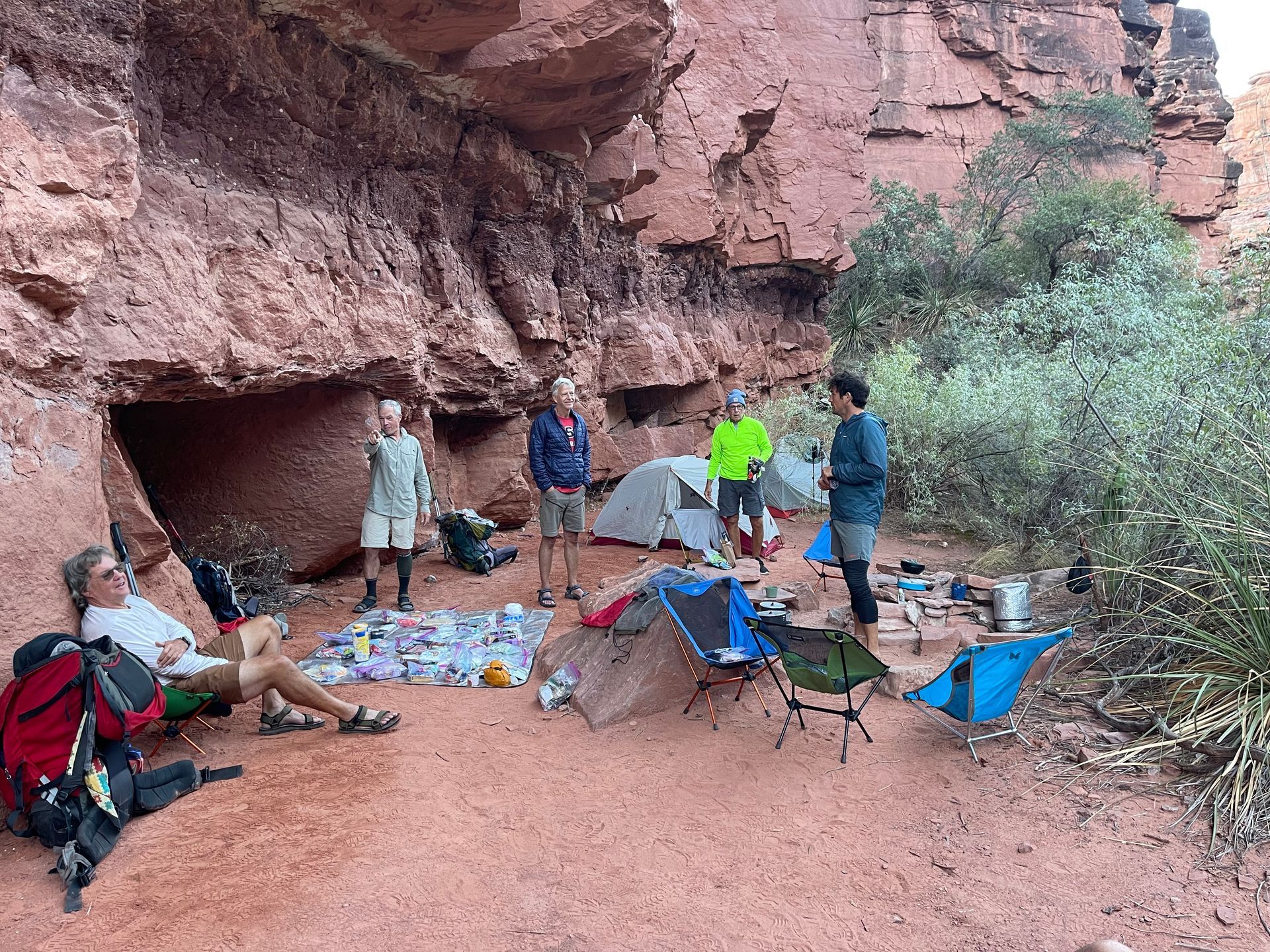 Campers relaxing under a red rock overhang. Tents, chairs, and gear are scattered about.