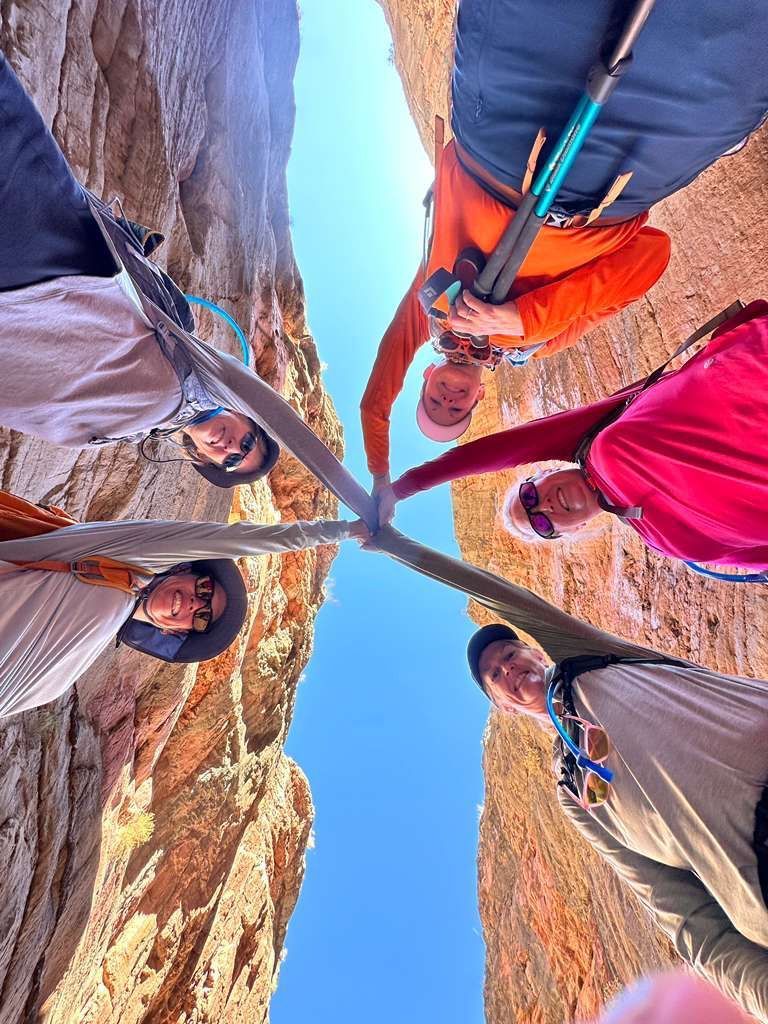 Five people with hiking poles look up, arms touching, between canyon walls. Blue sky.