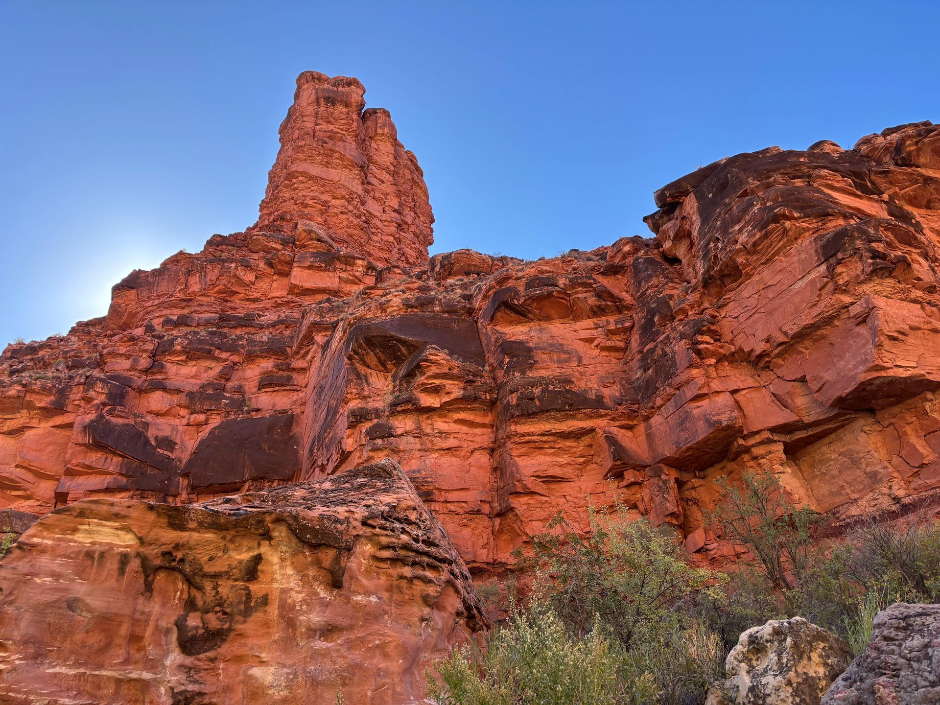 Red rock formation against a blue sky, with a tower-like structure and some foliage.