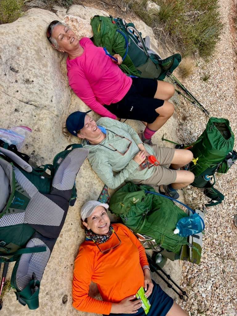 Three hikers rest on a rock. One in pink, one in green, one in orange. Backpacks and hiking poles nearby.