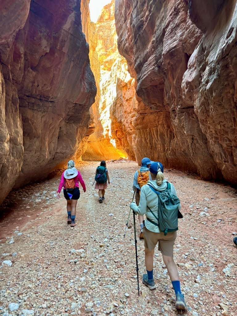 Hikers in a narrow red rock canyon, walking toward bright light.