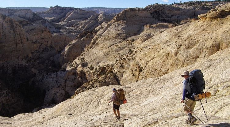 Two people with backpacks are hiking up a hill in the desert.