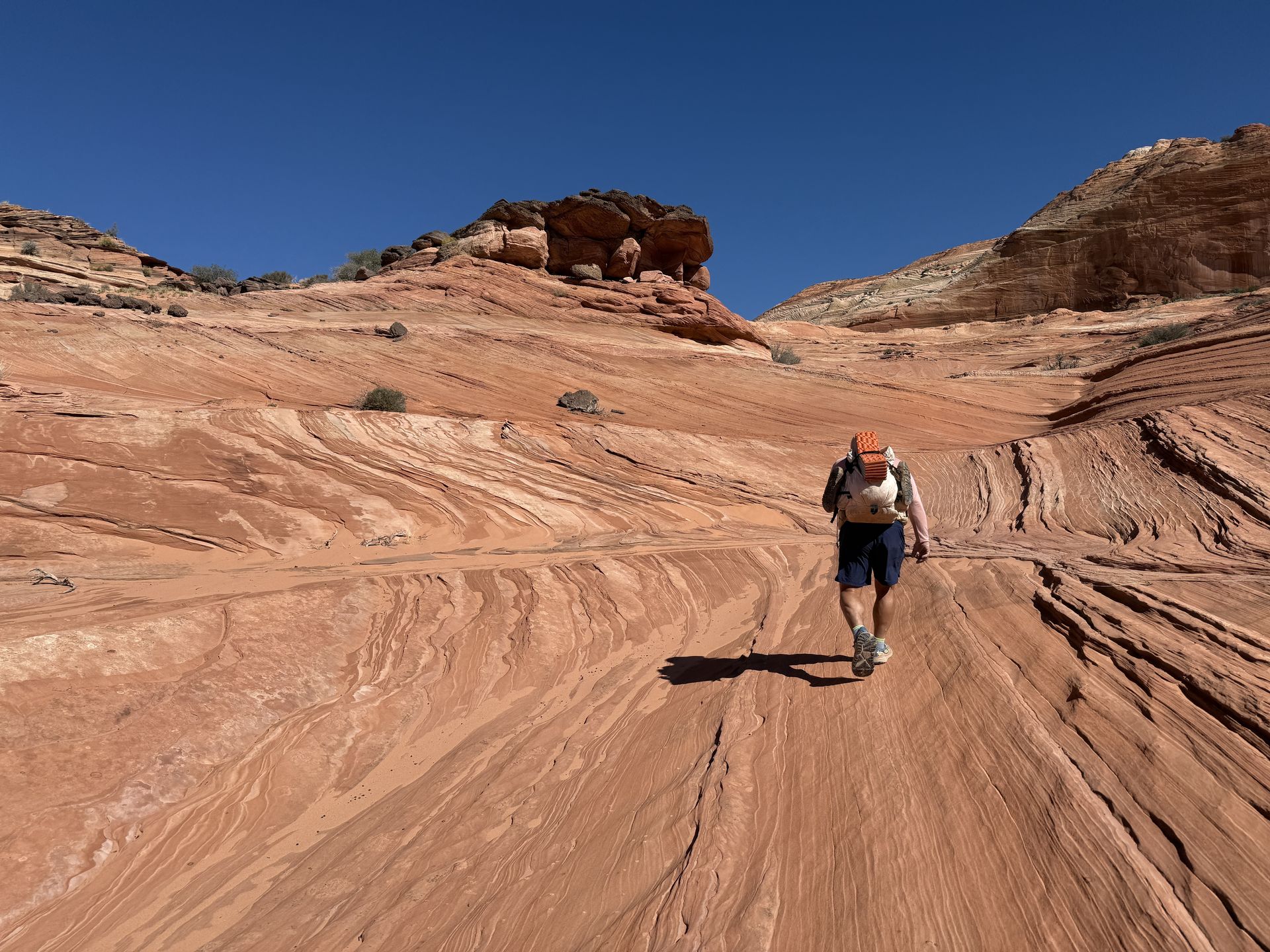 Person hikes up a red-rock slope under a clear blue sky, carrying a backpack.