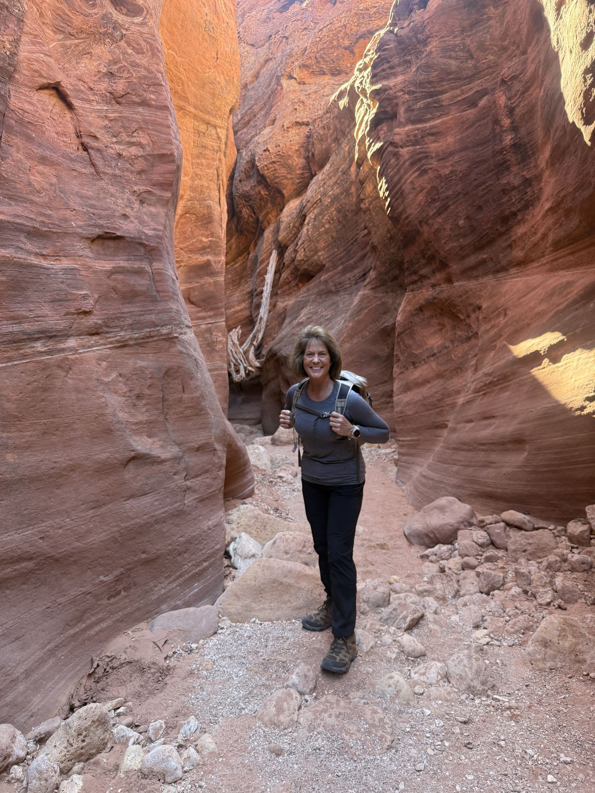Woman in canyon smiles, wearing backpack. Hiking between red rock walls.