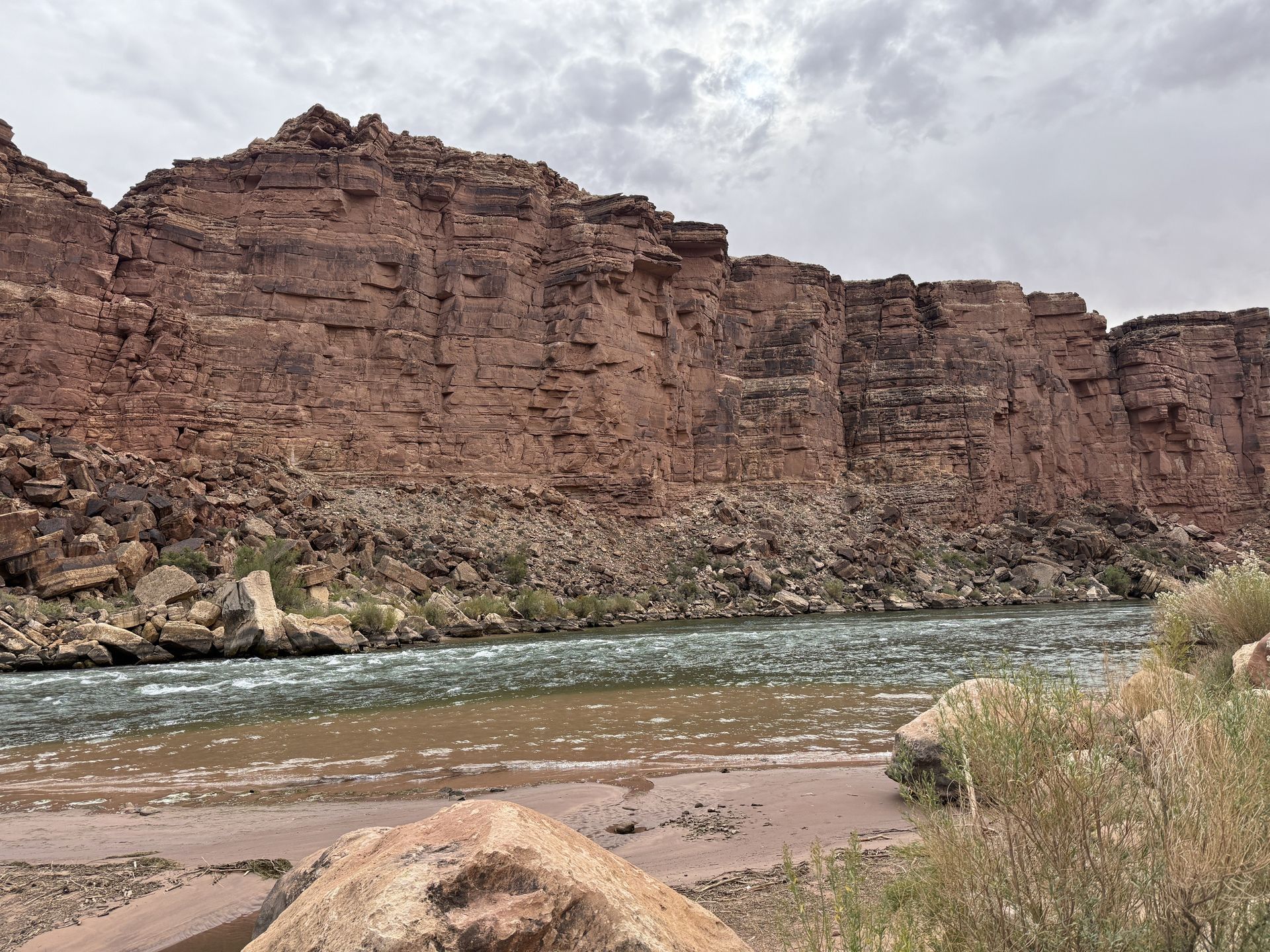 River flowing in front of a red rock canyon under a cloudy sky.