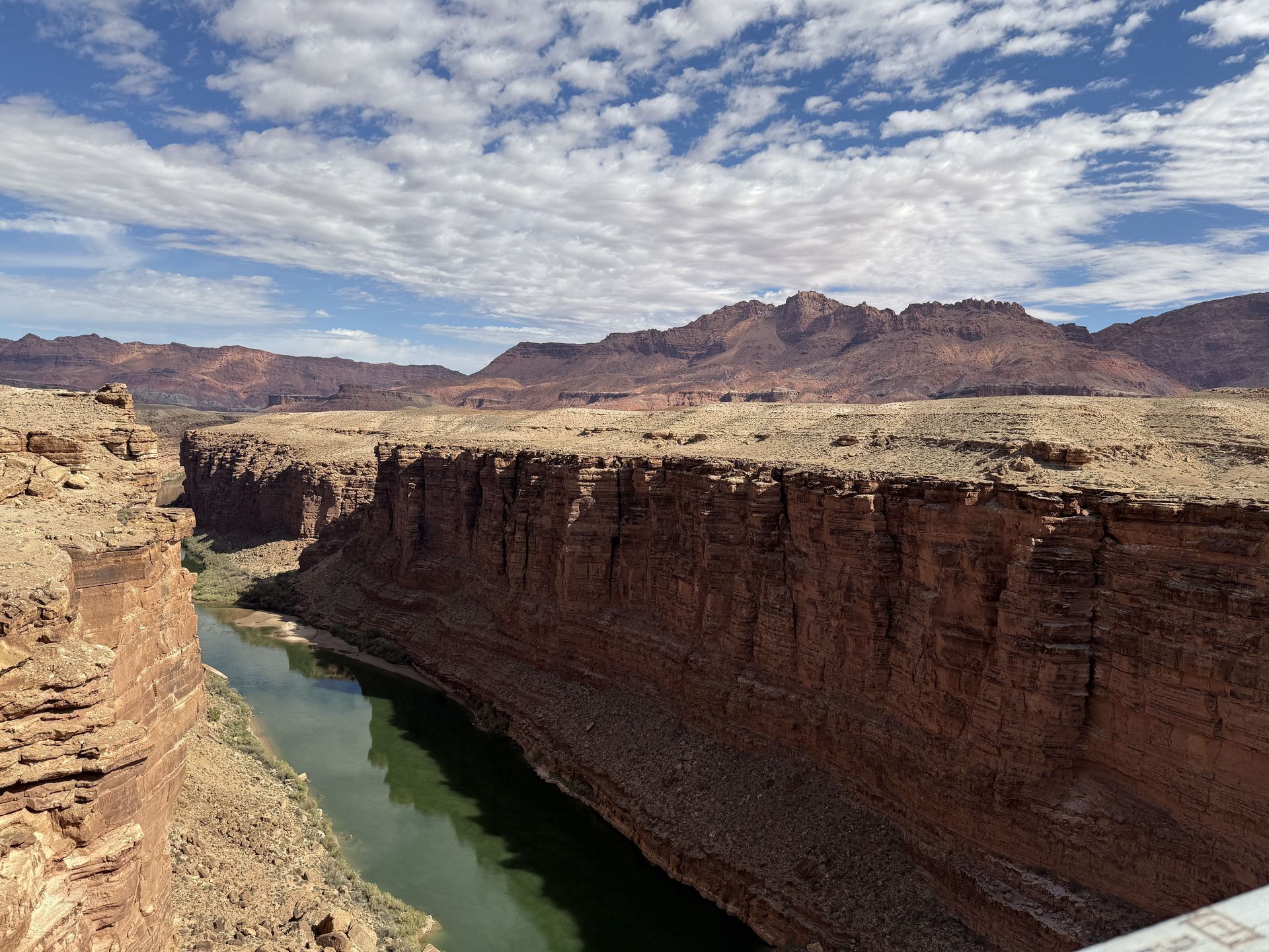 Canyon with a green river and reddish-brown rock formations under a cloudy blue sky.