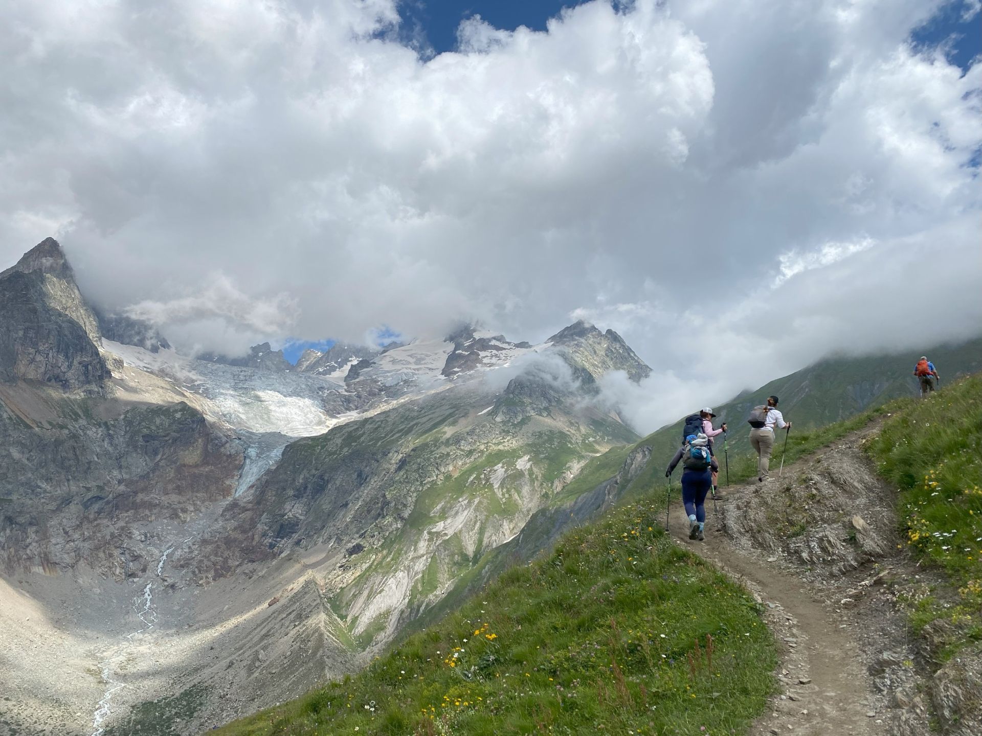 Hikers on a mountain trail, with a cloudy sky and snowy peaks in the background. Green grass and blue sky.