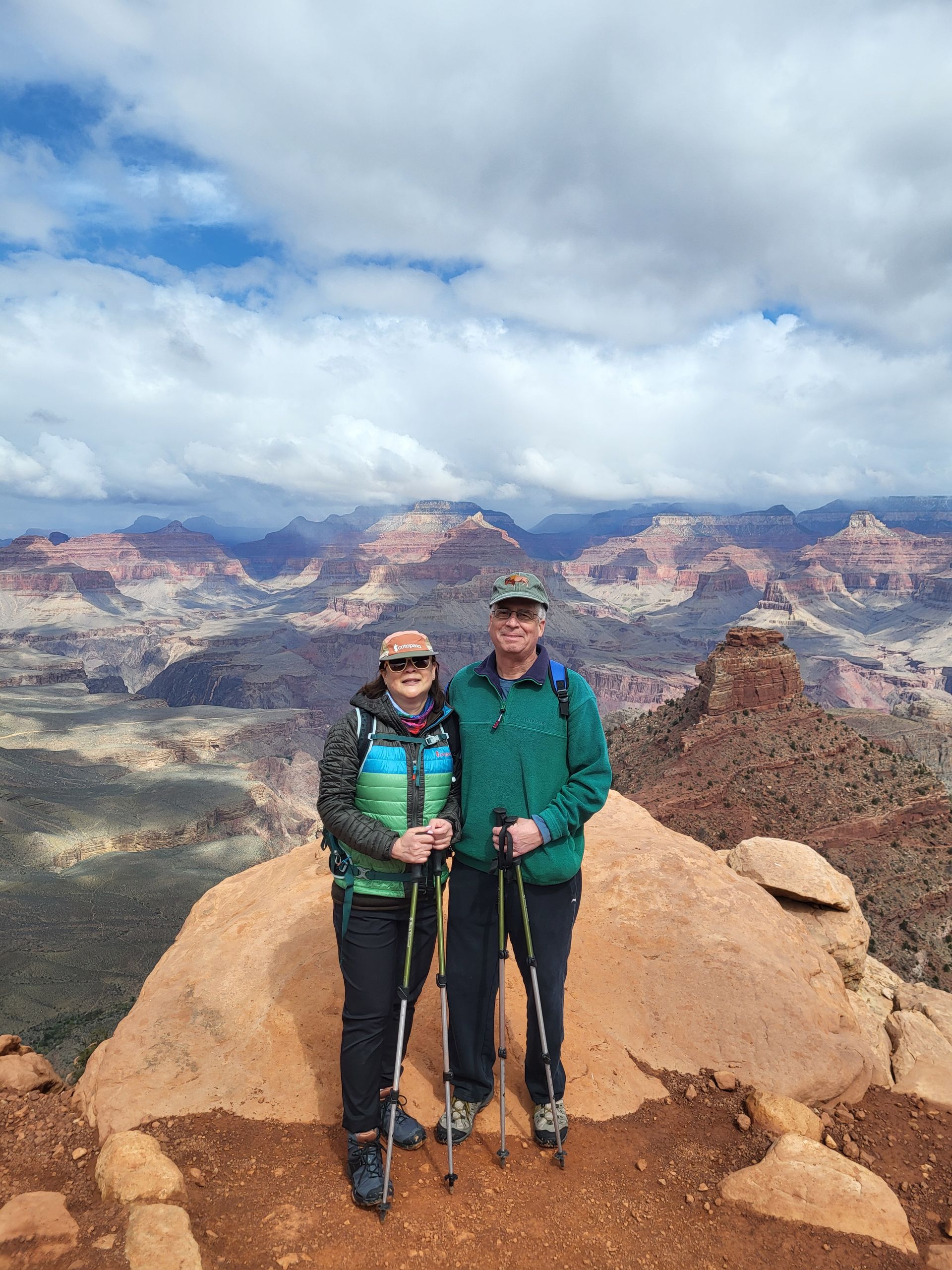 Couple with hiking poles poses on a red rock formation, vast canyon in background under cloudy sky.