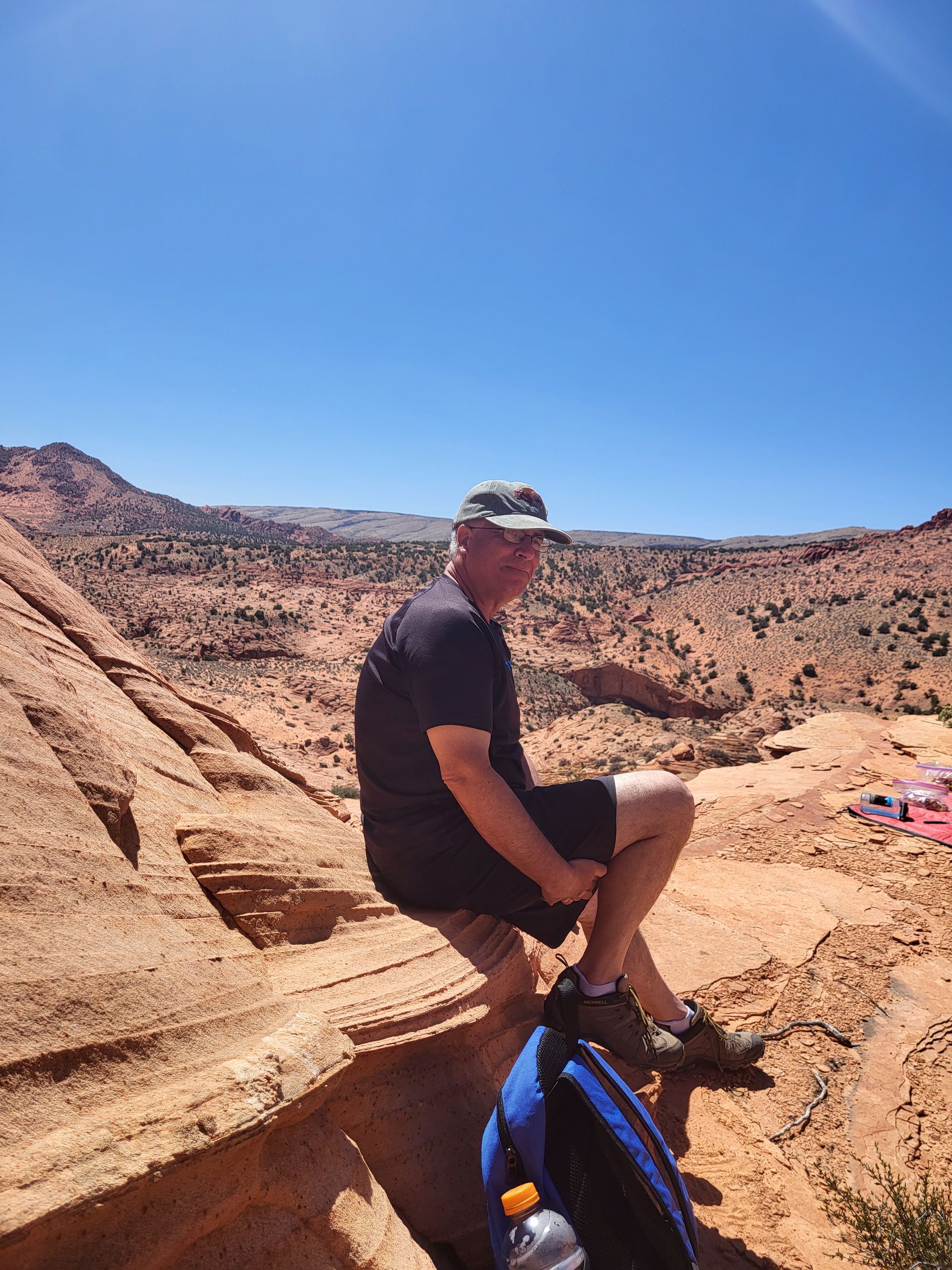 Man in hat sits on red rock ledge, overlooking desert landscape under blue sky.