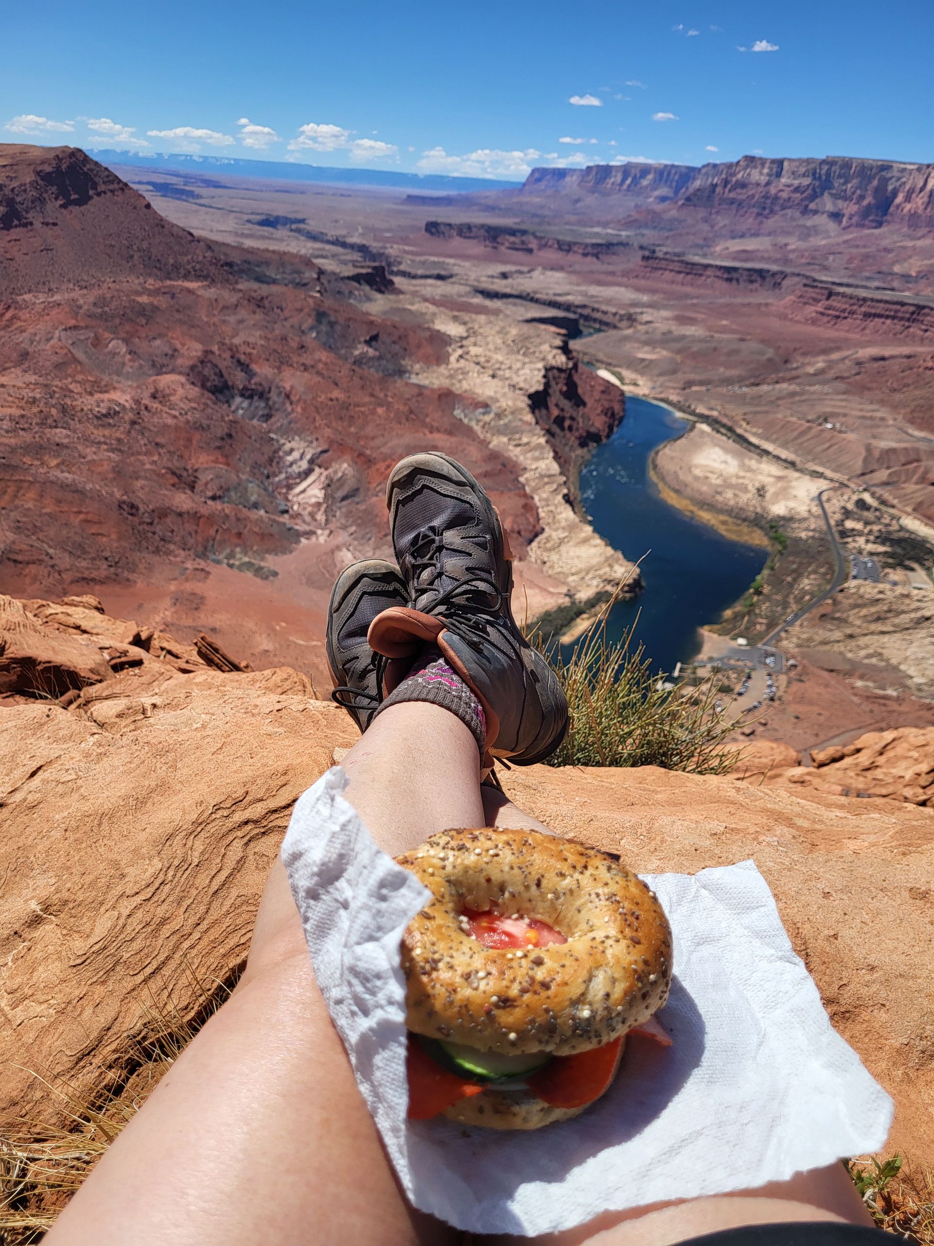 Person's legs with a bagel on a napkin, overlooking a canyon and water, blue sky.