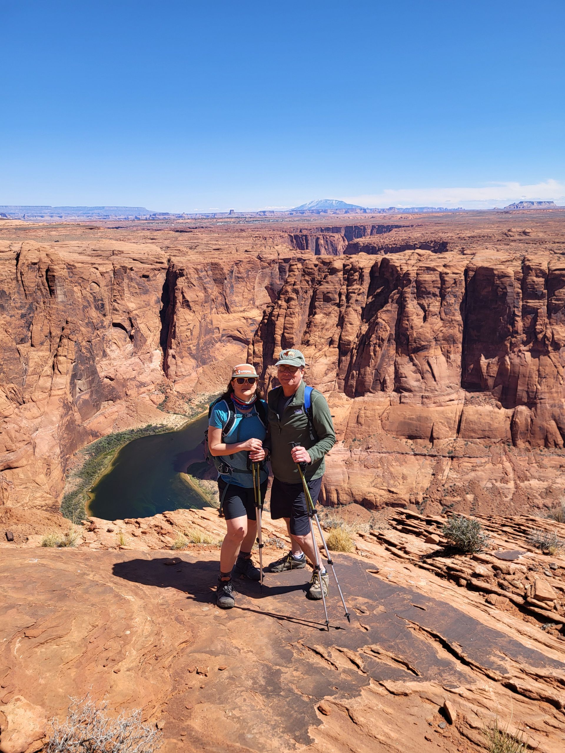 Two people pose on a red rock cliff overlooking a canyon with a lake, sunny blue sky.