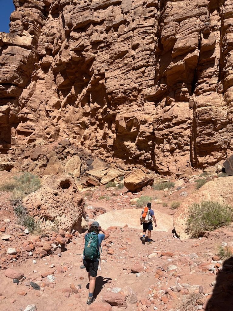 Two hikers with backpacks walk a rocky path through a canyon with red rock walls.