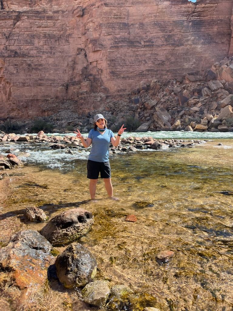 Person standing in shallow water, gesturing peace signs, against red canyon wall.