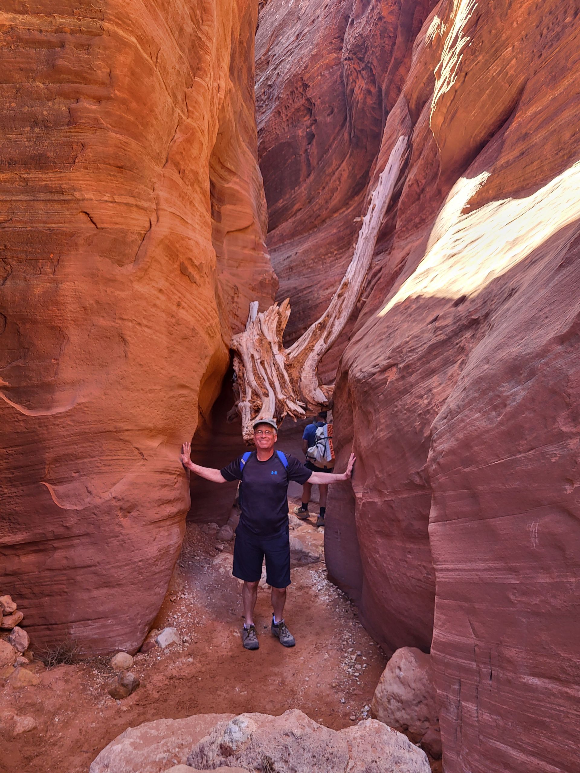 Man standing in a narrow slot canyon with arms outstretched. Red rock walls, sunlight.