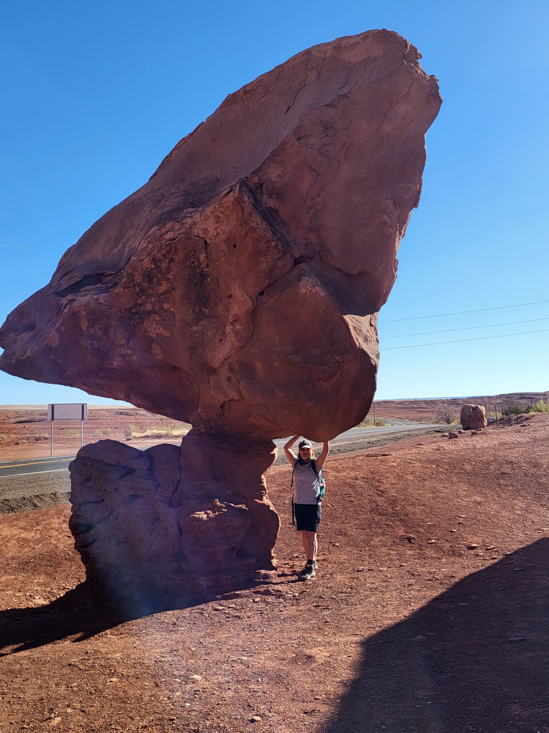 Person stands under large, precariously balanced rock formation in desert landscape.