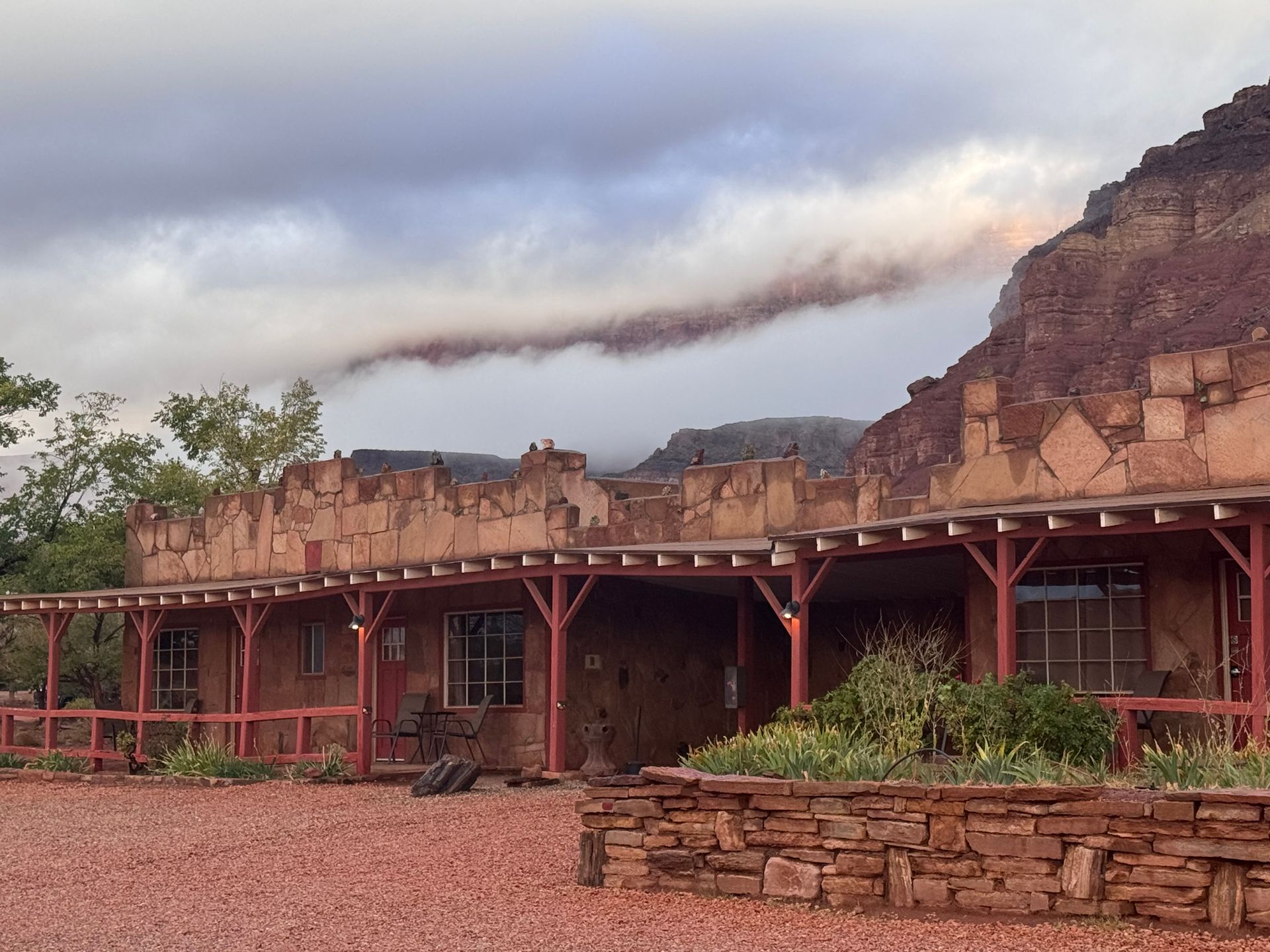 Red building with a wooden porch and rock facade, set against a red rock mountain, with low clouds.