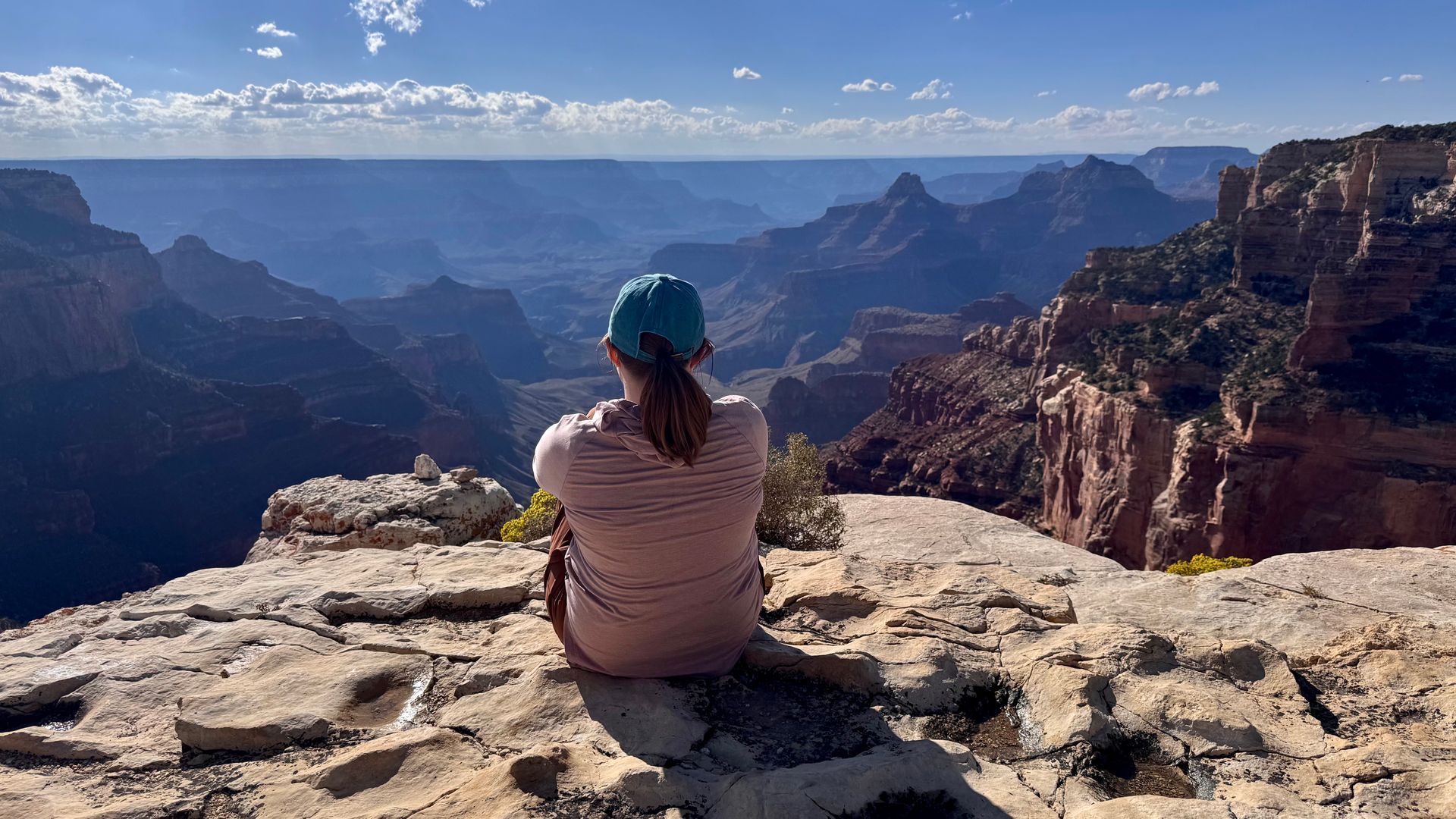Person sits on a cliff edge, looking at the Grand Canyon. Blue hat, beige top, sunny day.