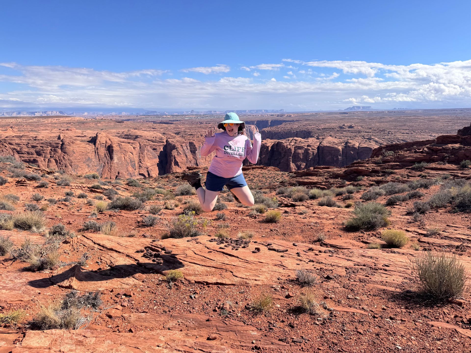 Woman jumping in front of a canyon. She wears a pink shirt, blue shorts, and a hat. Red rocks and blue sky.