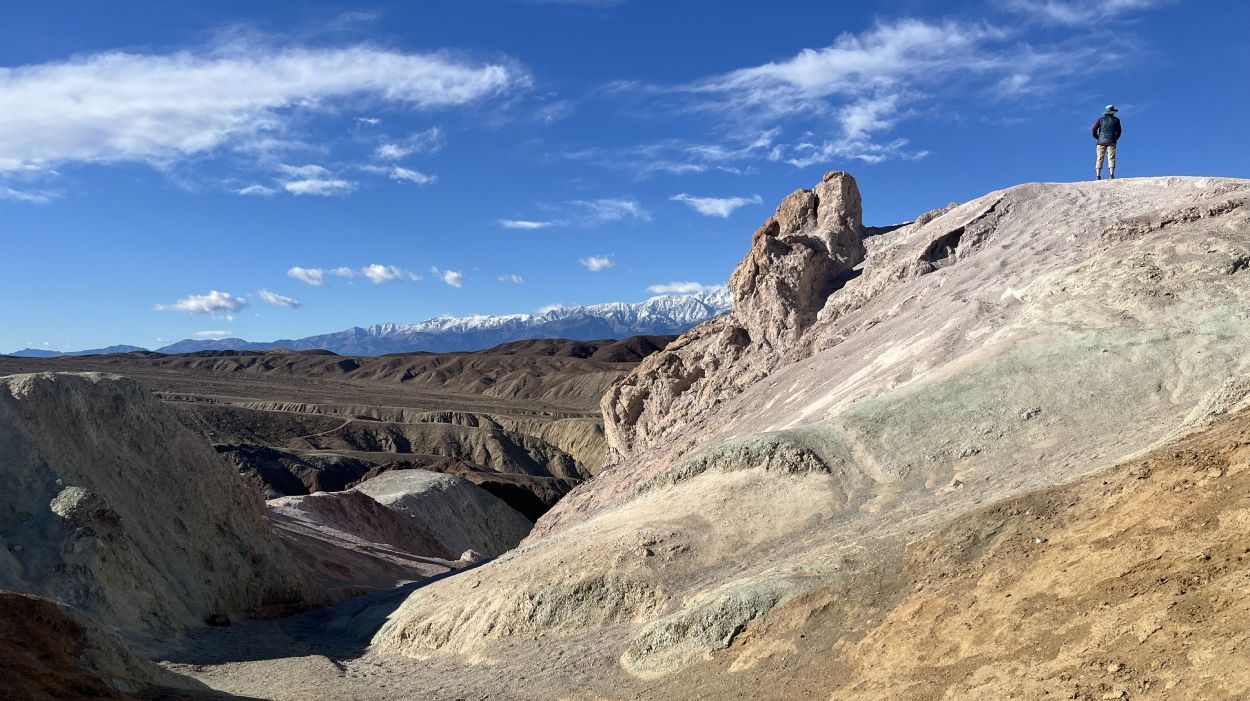 A person stands on a ridge overlooking a colorful, arid desert landscape with distant snow-capped mountains under a sky.