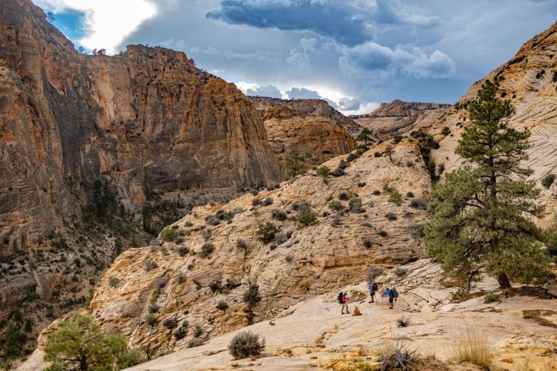 Hikers traverse a rocky, sunlit desert canyon landscape with dramatic, towering sandstone walls under a cloudy sky.