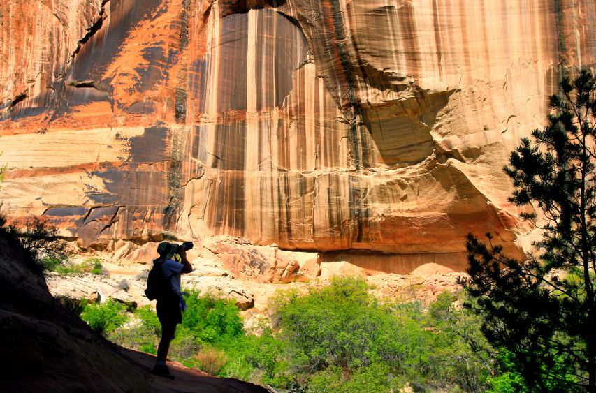 A photographer stands in a canyon, capturing a large natural stone arch against towering, striped sandstone walls.