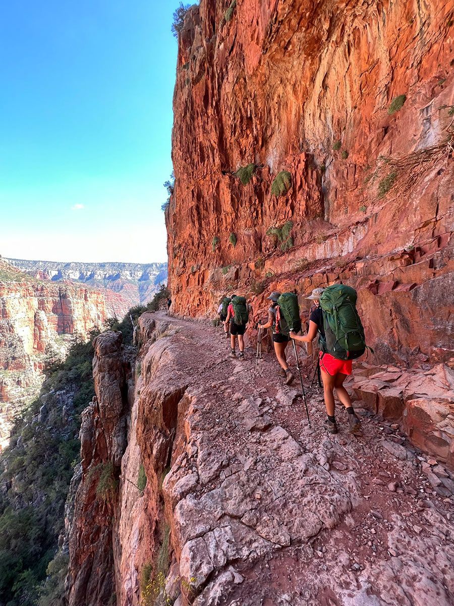 People with backpacks are hiking down a trail next to a cliff.