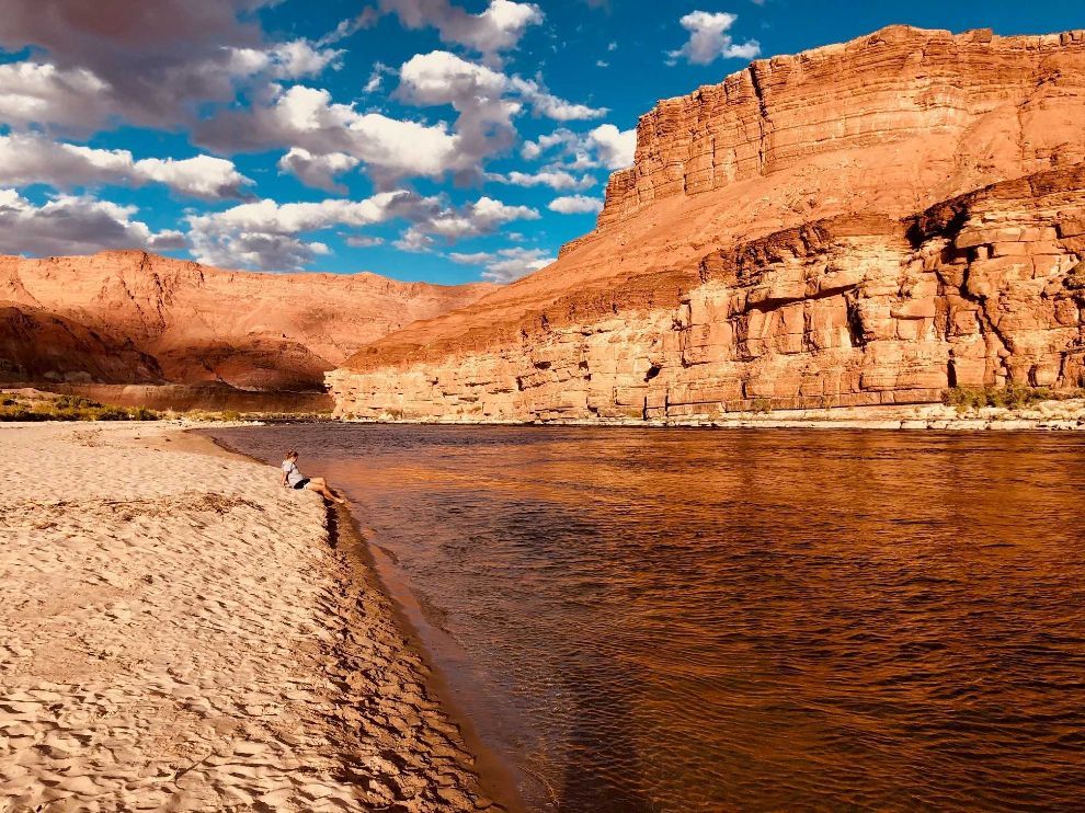 Sandy riverbank beside a red canyon under a blue sky with scattered clouds