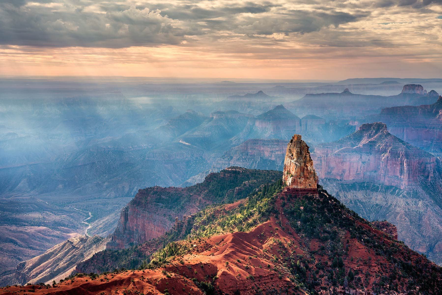 Canyon vista with a rocky pinnacle; hazy blues, greens, and reds under a cloudy sky.