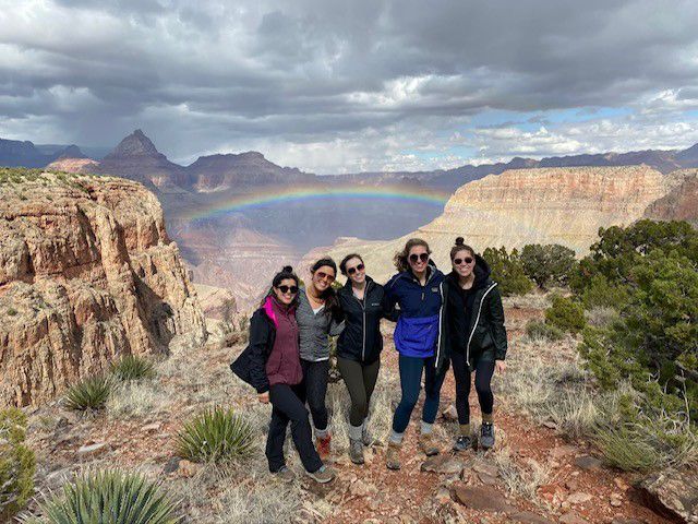 Five people pose on a cliff overlooking the Grand Canyon with a rainbow.