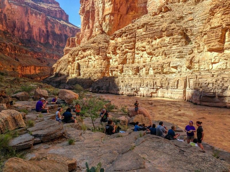 A group of people are sitting on rocks near a river in a canyon.