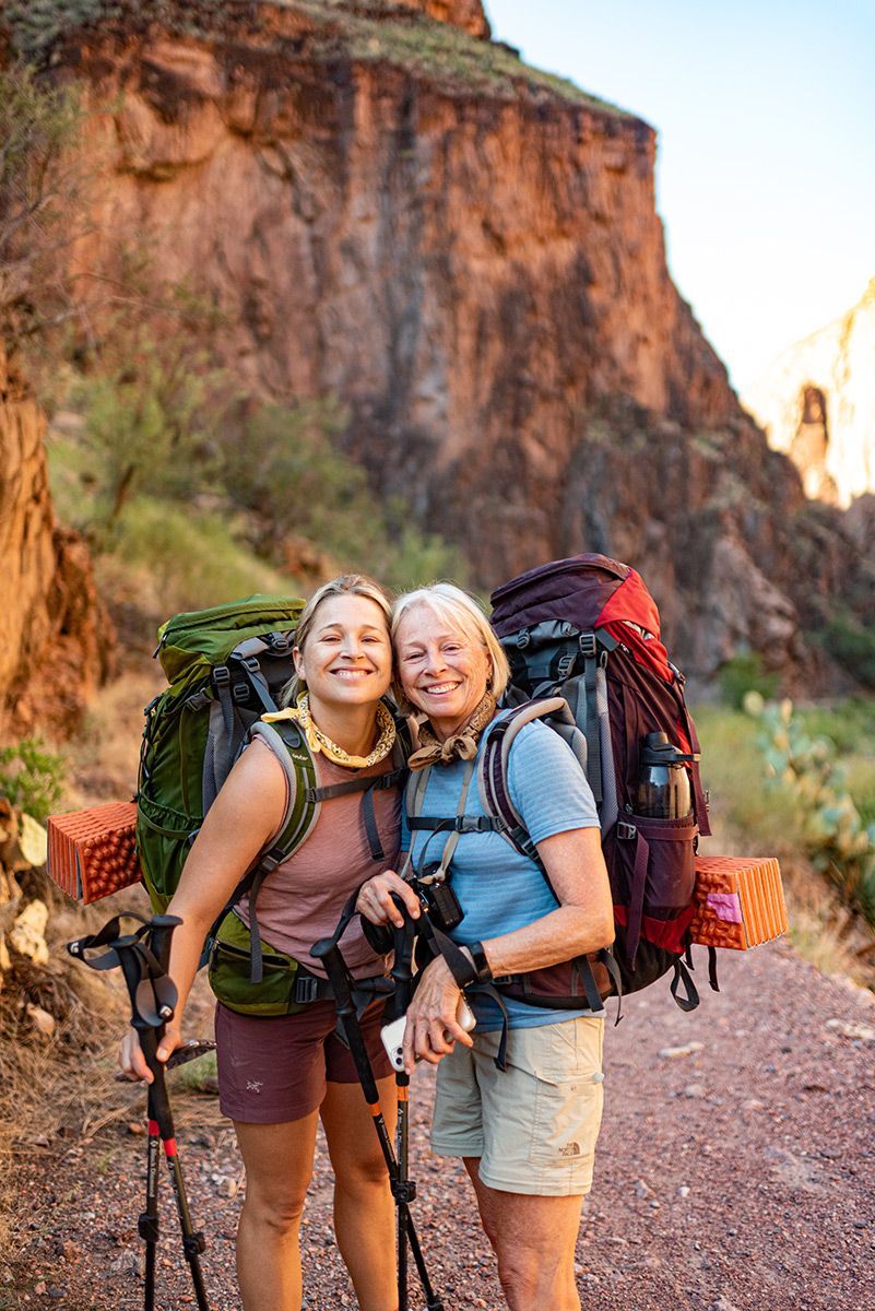 Two women with backpacks are standing next to each other on a dirt road.