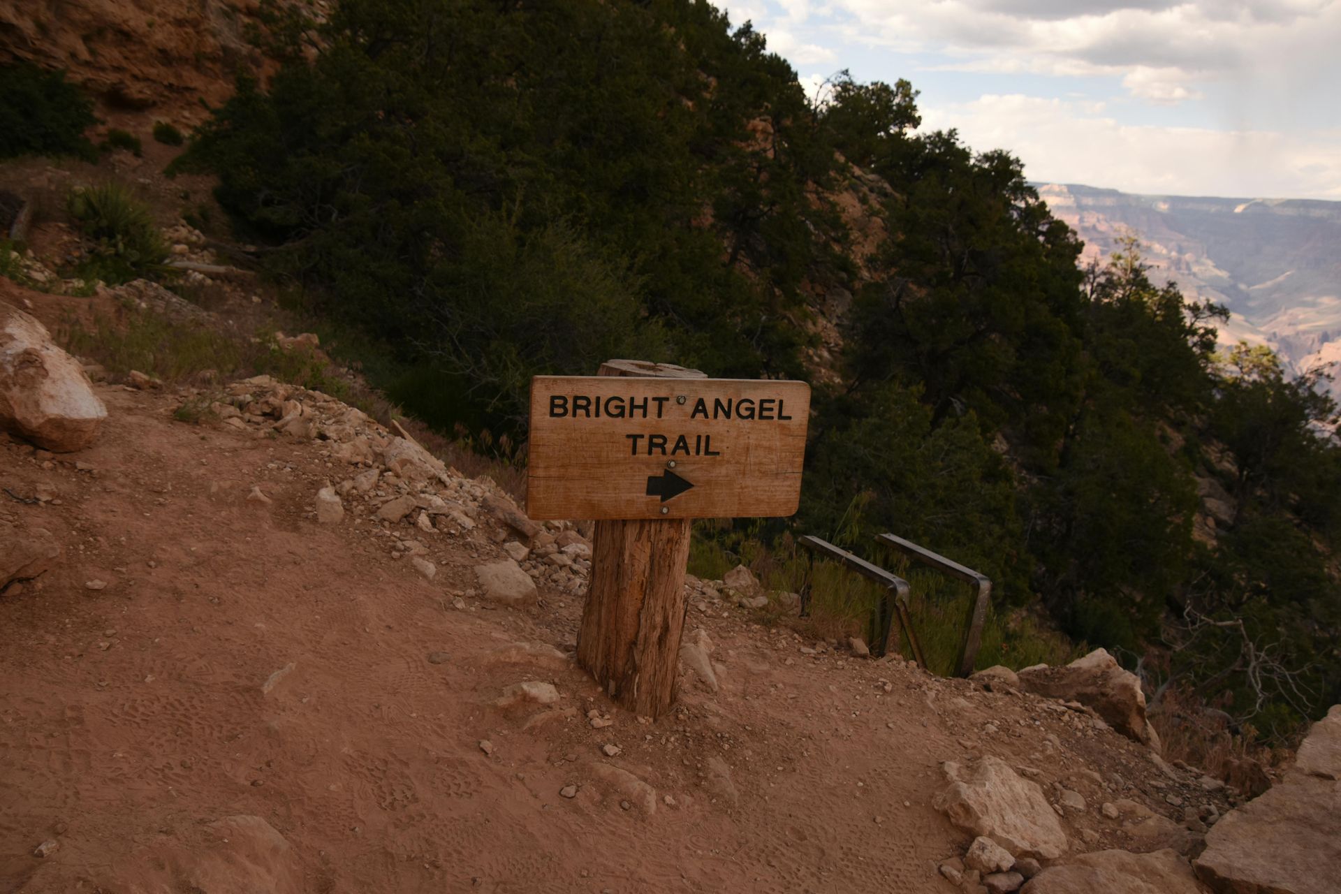 Bright Angel Trail sign on a rocky canyon path with trees and distant cliffs