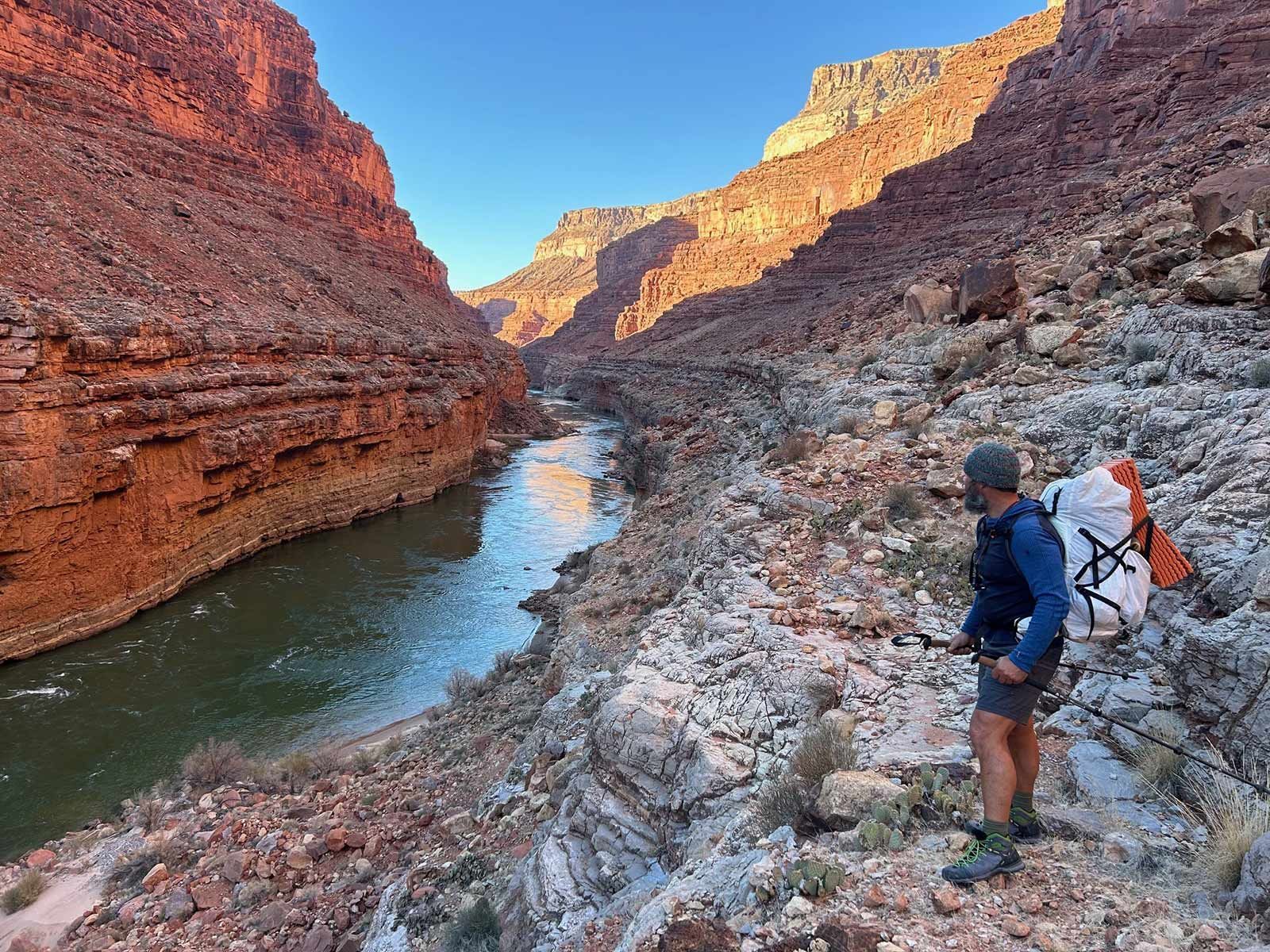 A man with a backpack is standing on a rocky cliff overlooking a river.