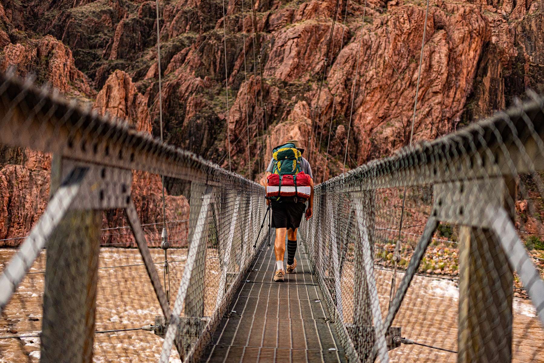 A person with a backpack is walking across a suspension bridge over a river.