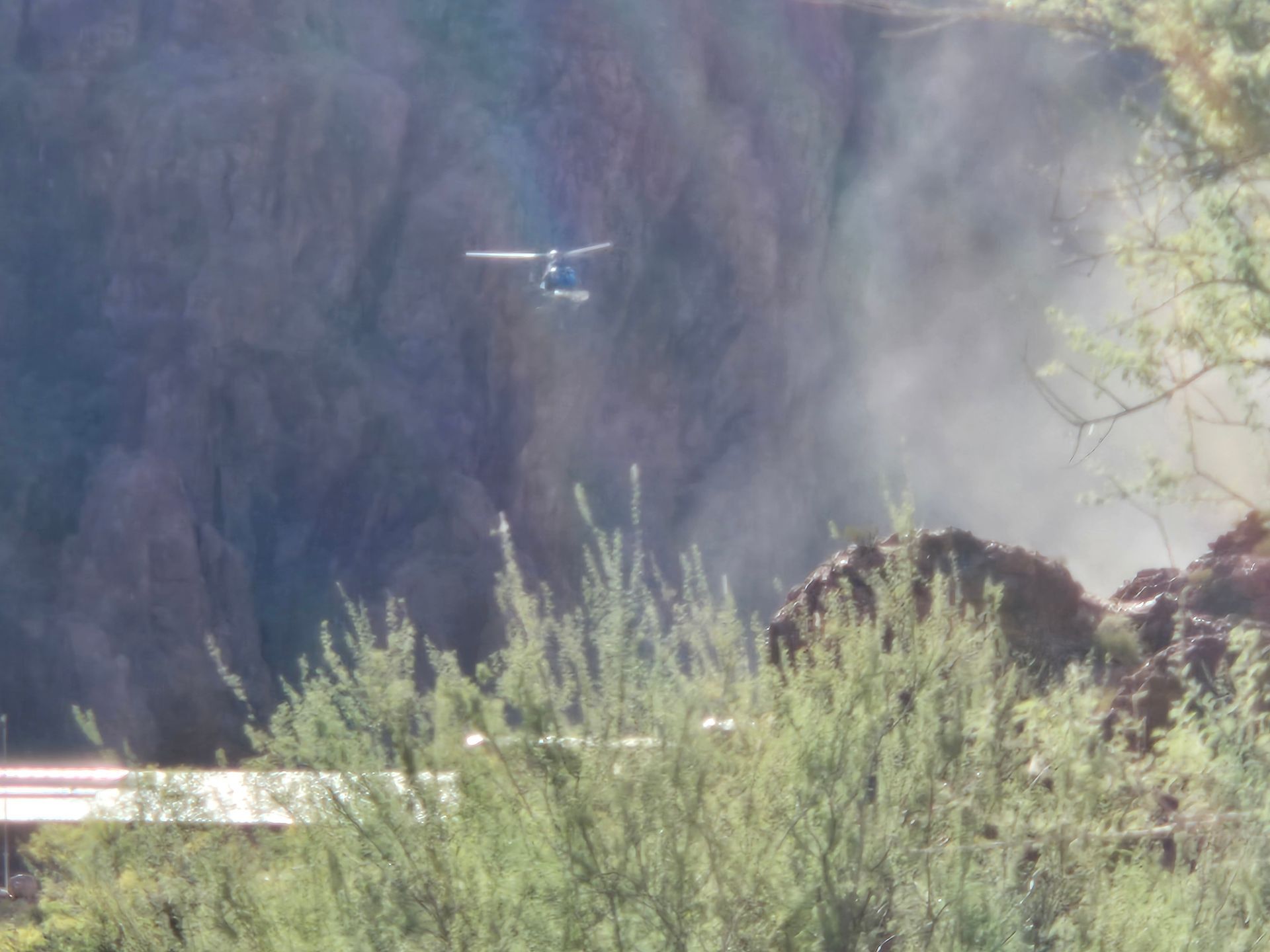 Helicopter flying near a rocky cliffside, with smoke visible. Green bushes in the foreground.